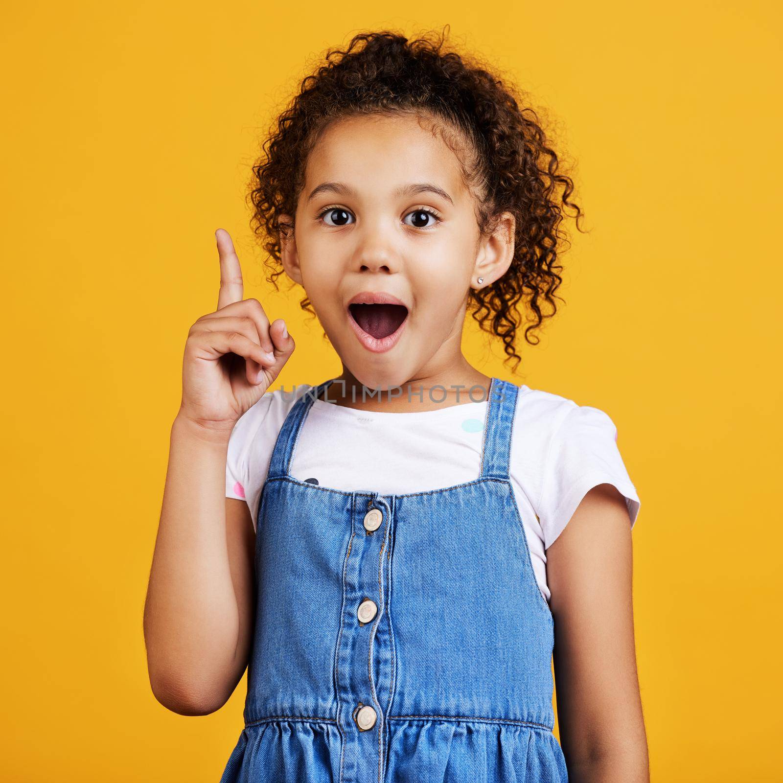 Studio portrait mixed race girl pointing upwards towards copyspace isolated against a yellow background. Cute hispanic child posing inside. Happy and cute kid showing or endorsing a company or product.