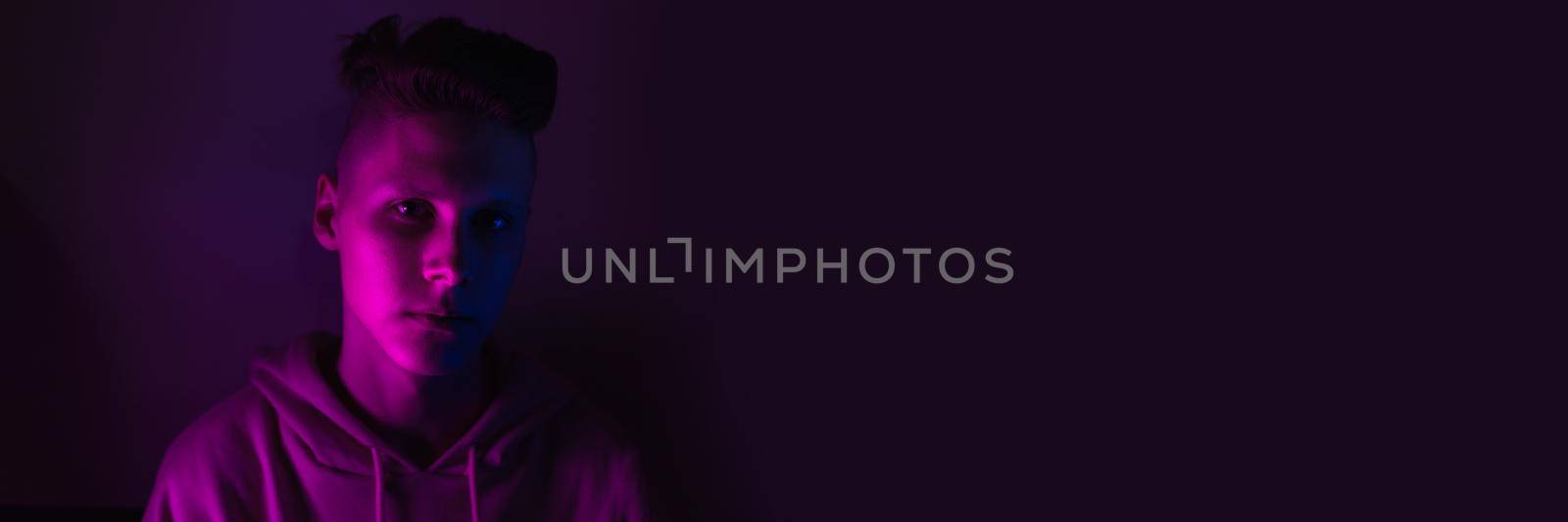 Young guy in a neon light. A teenager in a pink hoodie in trendy lighting of pink, purple and blue. Web banner.