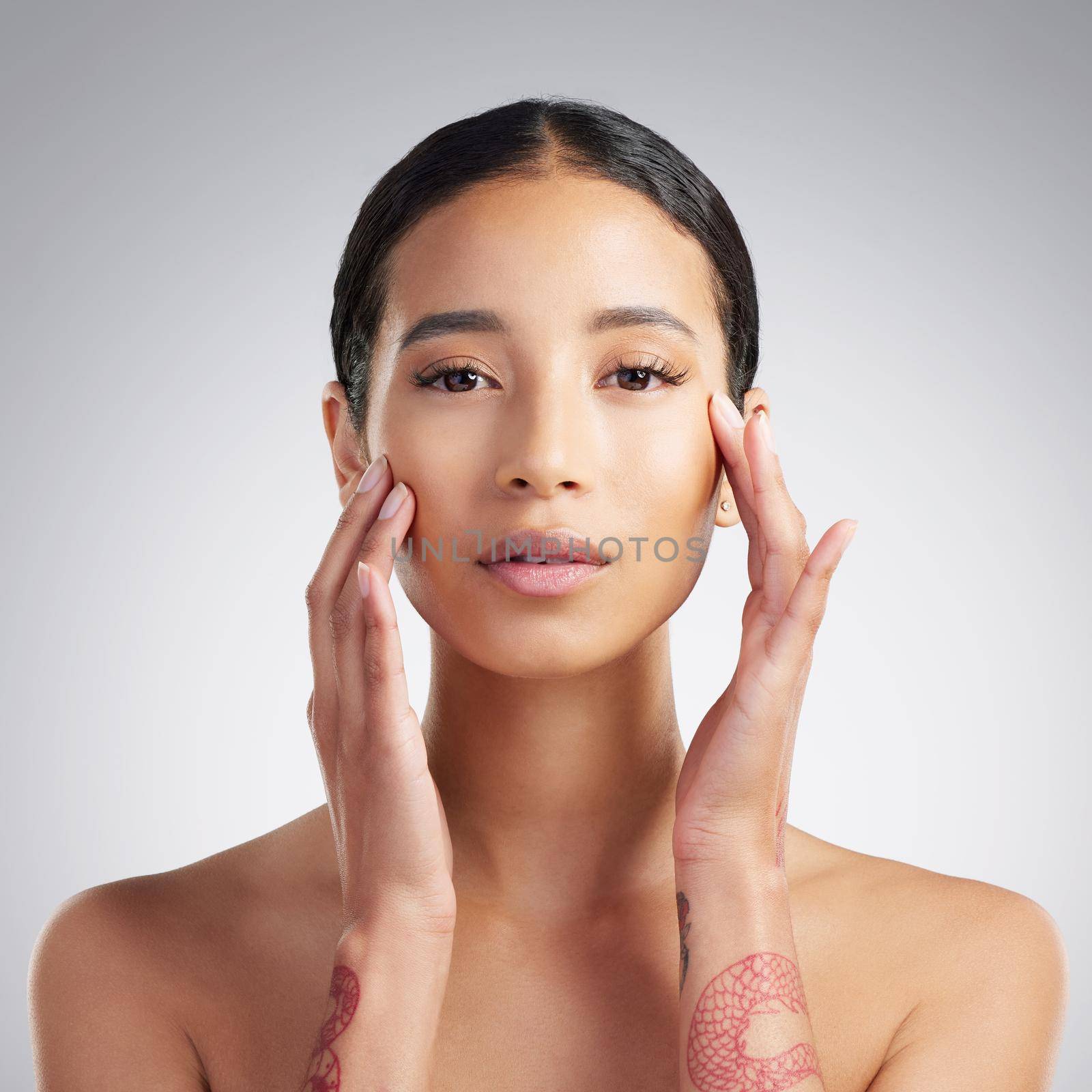 Portrait of a beautiful mixed race woman touching smooth soft skin in a studio. Hispanic model with healthy natural glowing skin looking confident against grey copyspace while doing routine skincare by YuriArcurs