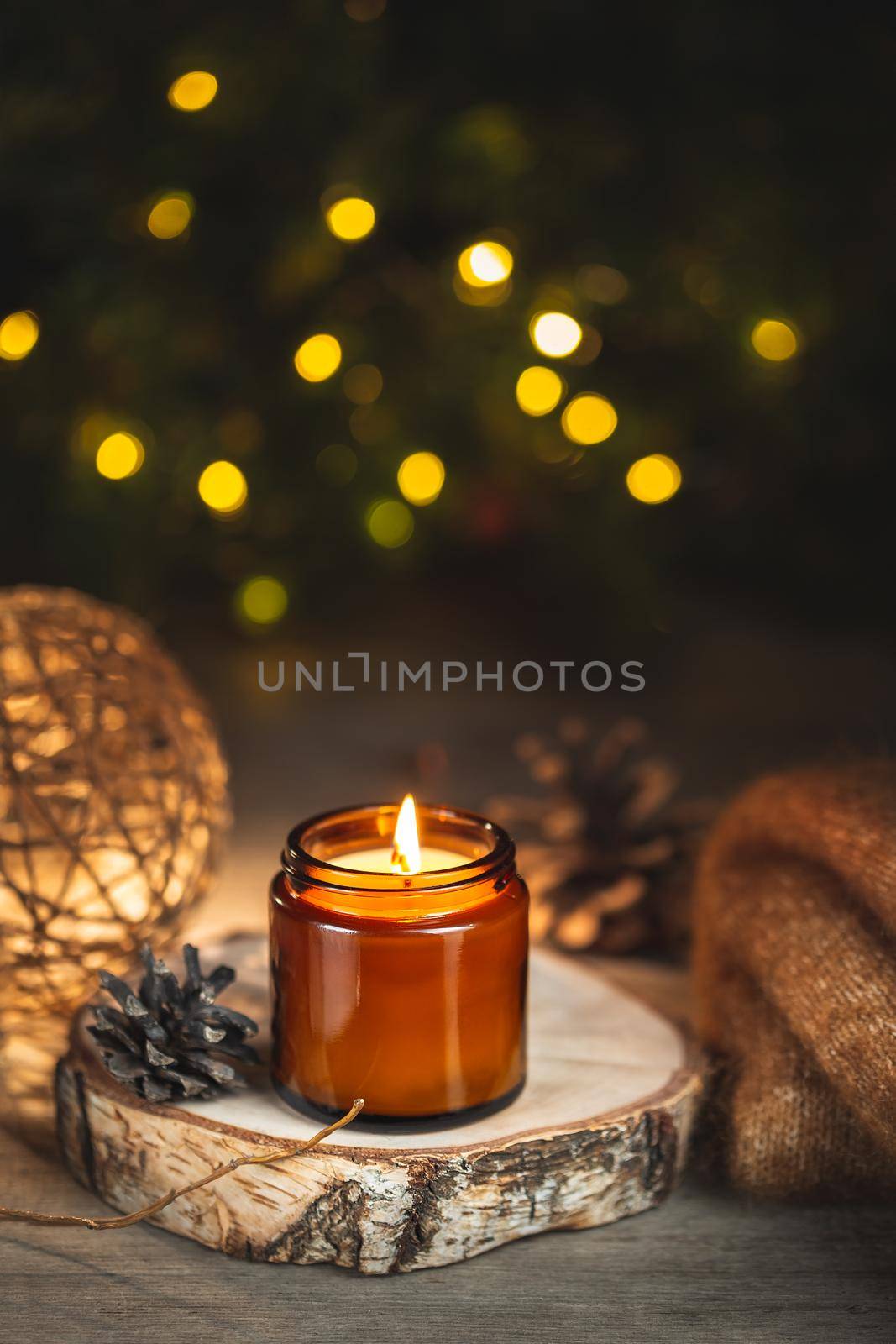 Cozy winter evening card with burning aroma candle in a dark glass jar over blurred bokeh lights on the background. Soft focus, copy space for text. Christmas holiday at home.