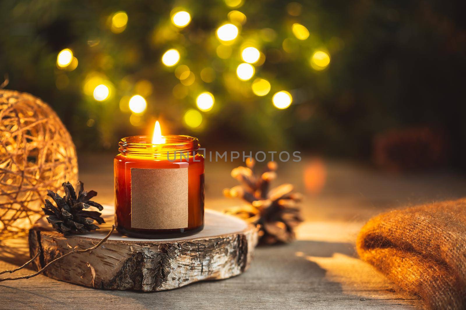 Cozy winter evening card with burning aroma, candle in a dark glass jar with empty label mock up. Soft focus, copy space for text. Blurred bokeh lights on the background. Christmas holiday at home.