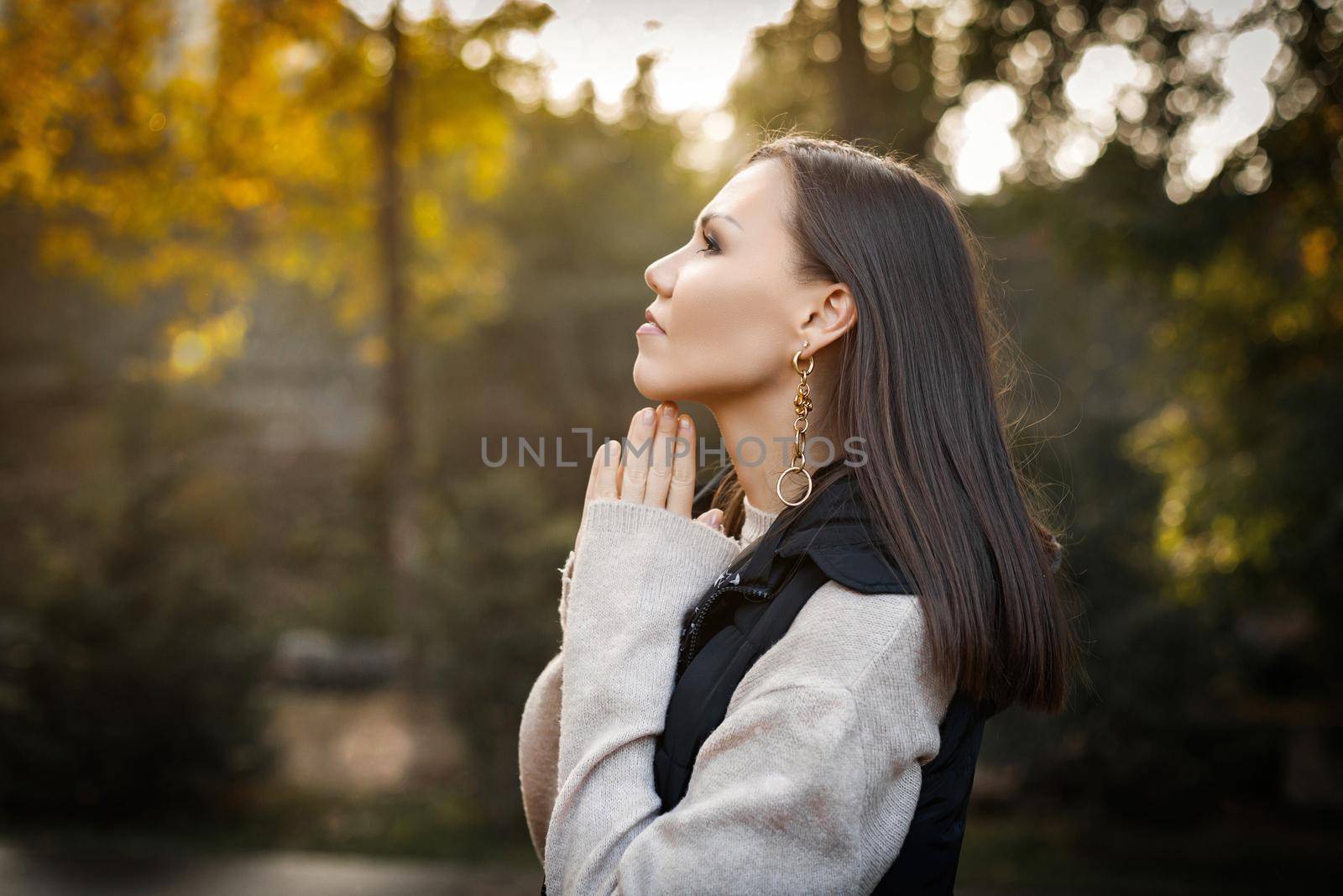 young serious pensive woman with hands near her face in autumn in a park outdoors in a warm beige jacket and sleeveless jacket.