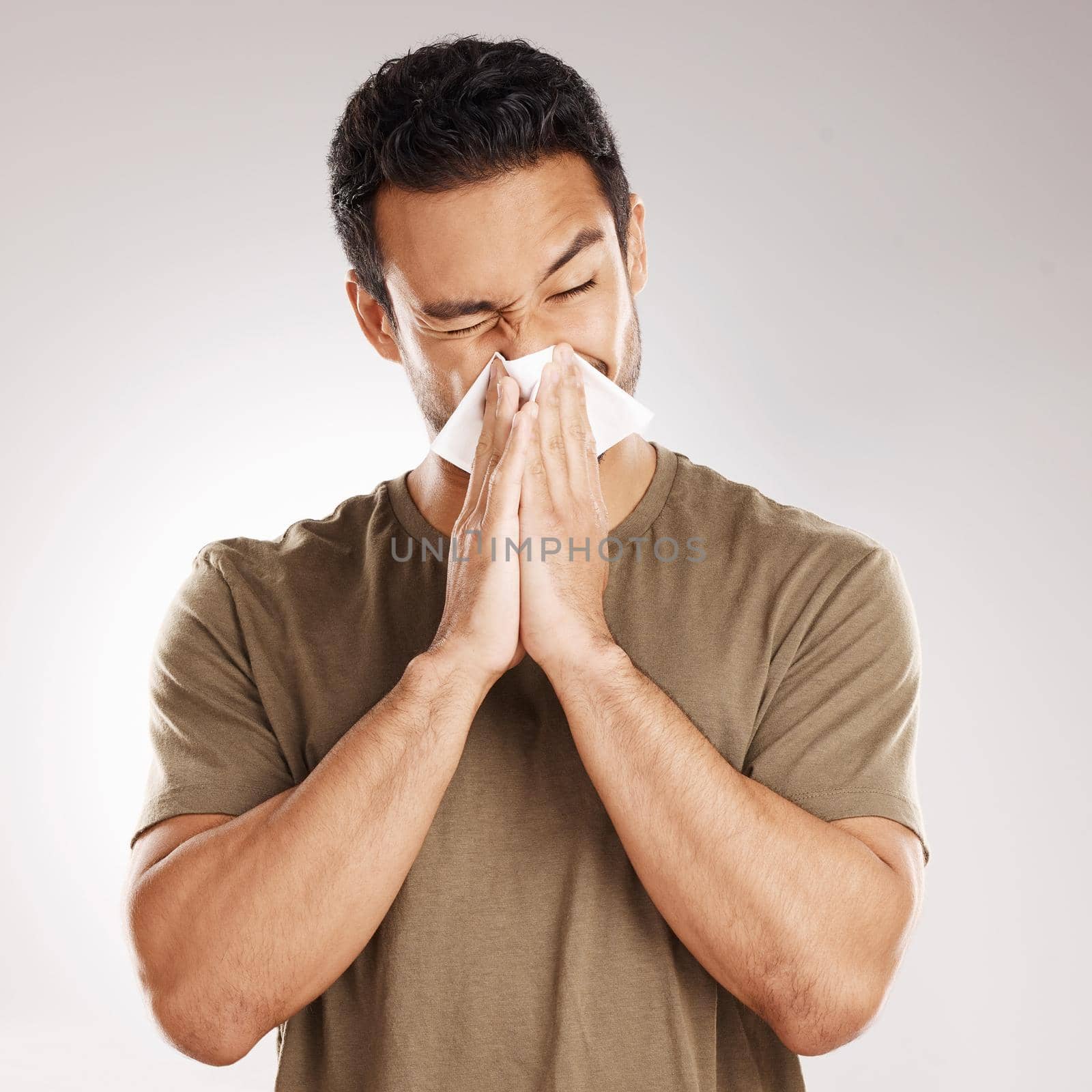 Handsome young mixed race man blowing his nose while standing in studio isolated against a grey background. Hispanic male suffering from cold, flu, sinus, hayfever or corona and using a facial tissue by YuriArcurs