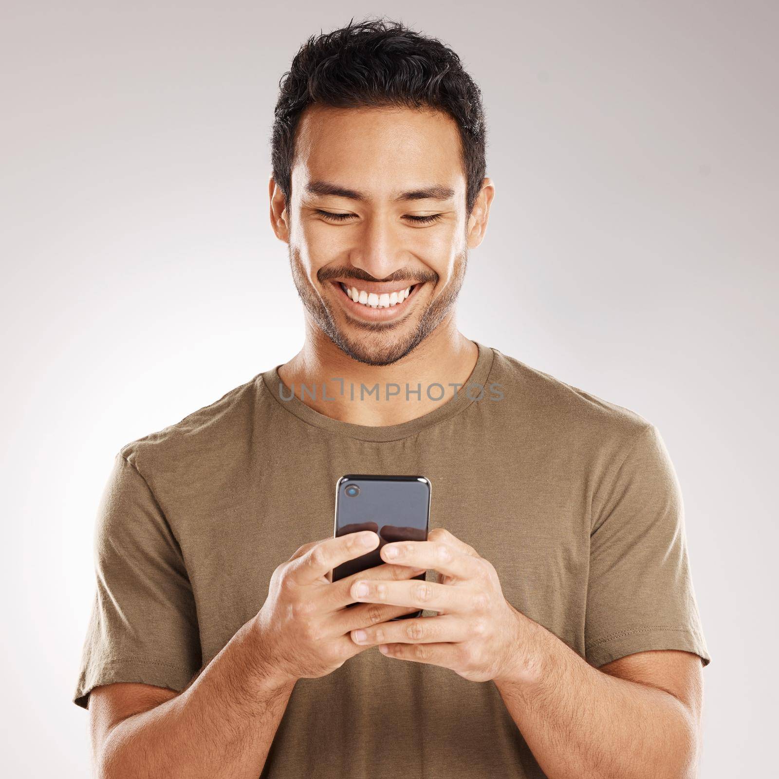 Handsome young mixed race man using his phone while standing in studio isolated against a grey background. Hispanic male sending a text message, using the internet online or browsing social media by YuriArcurs