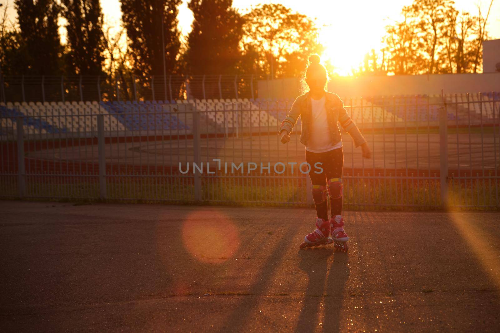 Little pretty happy funny girl on roller skates at stadium at sunset, learning to roller skate outdoors. Outdoor activity for children. Active sport for preschool kid. sun glare, selective focus