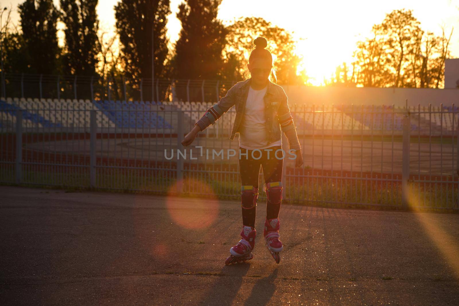 Little pretty happy funny girl on roller skates at stadium at sunset, learning to roller skate outdoors. Outdoor activity for children. Active sport for preschool kid. sun glare, selective focus