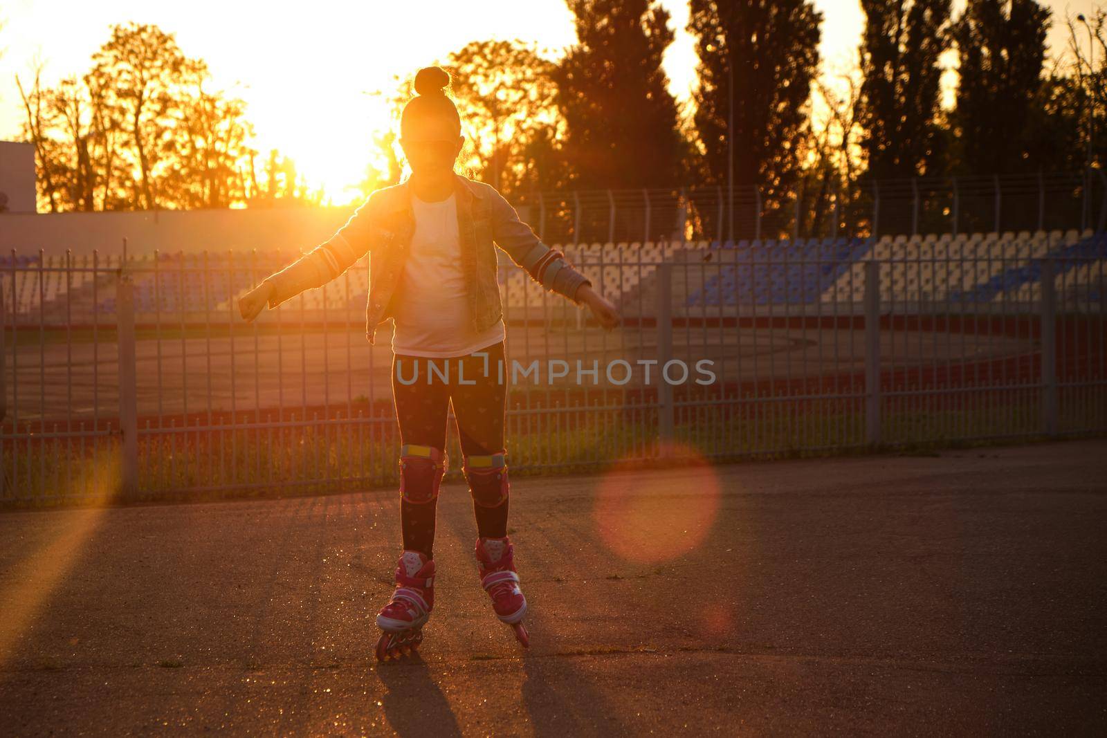 Little pretty happy funny girl on roller skates at stadium at sunset, learning to roller skate outdoors. Outdoor activity for children. Active sport for preschool kid. sun glare, selective focus