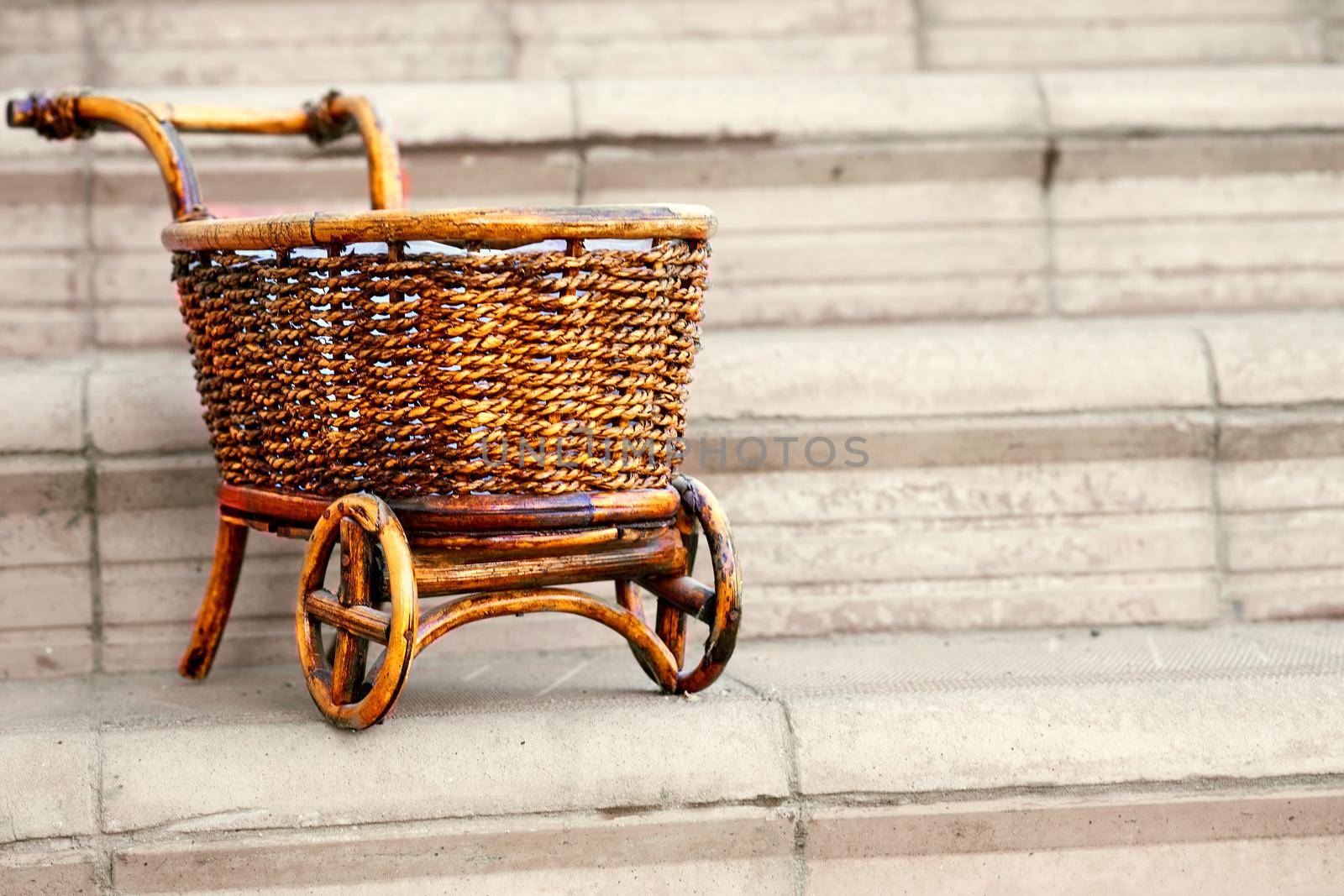 a strong open vehicle with two or four wheels, typically used for carrying loads and pulled by a horse. Wooden wicker brown cart on pale gray steps.