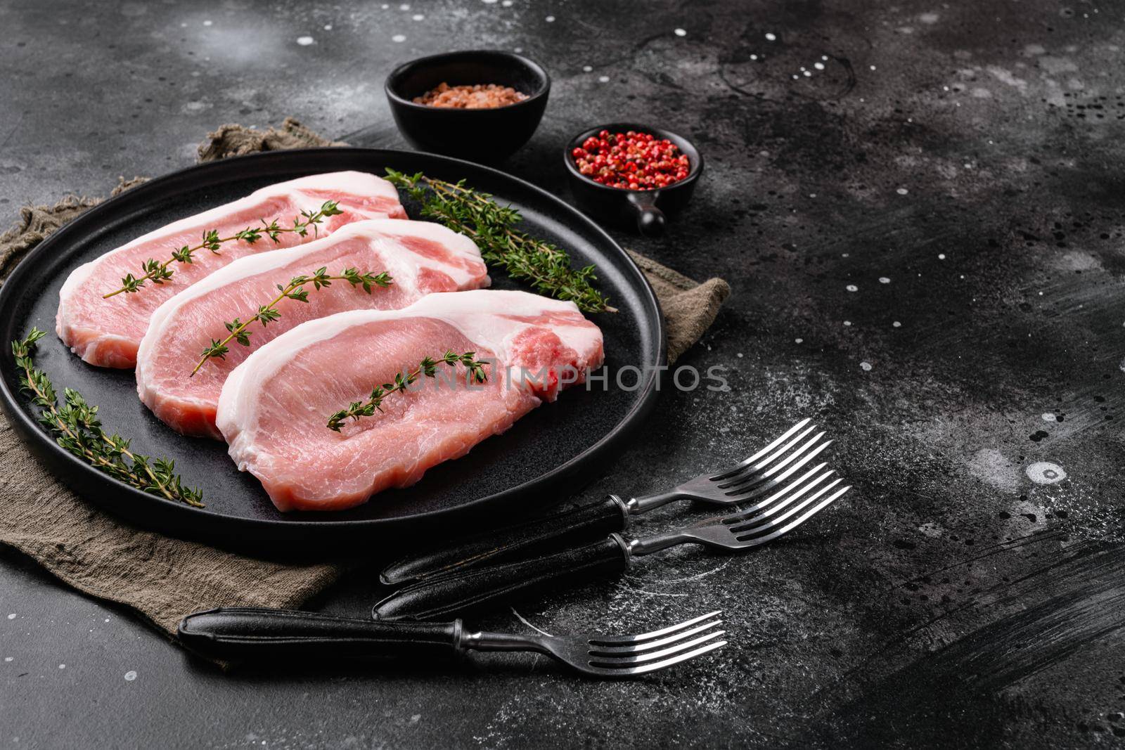 Raw pork steak ready for cooking with herbs set, on black dark stone table background, with copy space for text