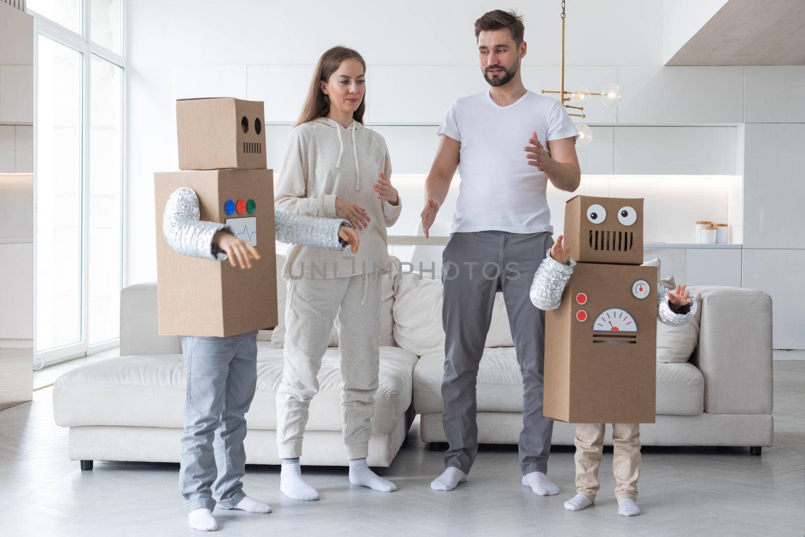 Happy family of parents and two children playing dancing like robots at home, children wearing handmade moving box costume of cardboard