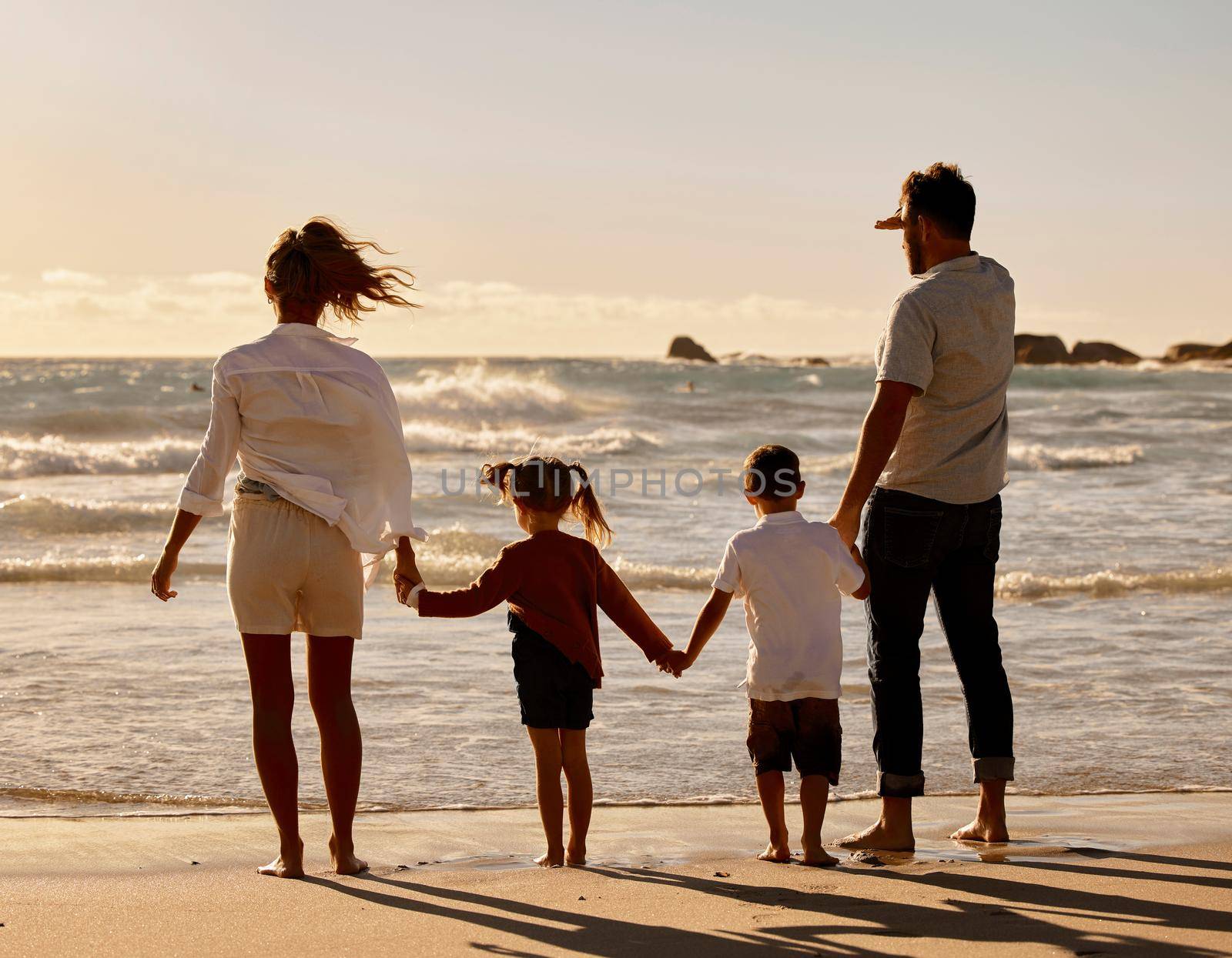 Back of caucasian parents with son and daughter enjoying free time on a beach. Little boy and girl holding hands and bonding with their mother and father on the weekend. Family watching the sunset.