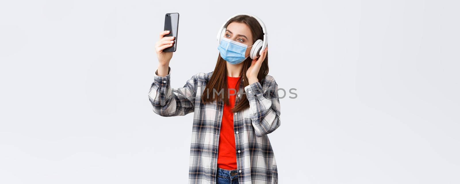 Social distancing, leisure and lifestyle on covid-19 outbreak, coronavirus concept. Woman in headphones and medical mask listening music, taking selfie on mobile phone using filters.