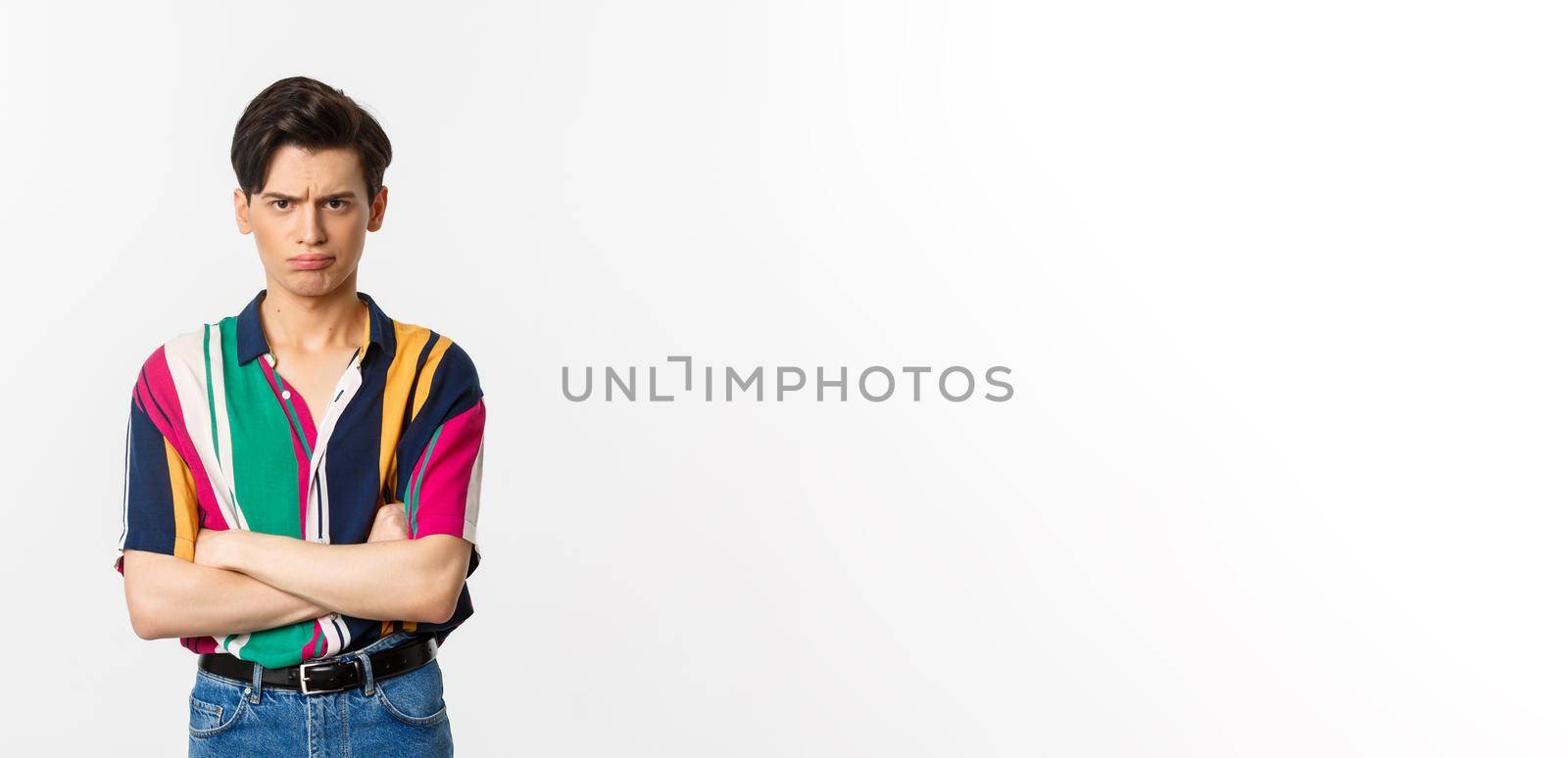 Offended and angry young gay man sulking, cross arms chest and looking judgemental at camera, standing over white background.