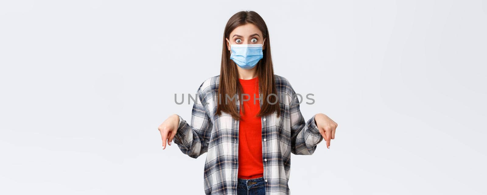 Coronavirus outbreak, leisure on quarantine, social distancing and emotions concept. Shocked and impressed young woman in medical mask and casual outfit, pointing fingers down, stare camera.