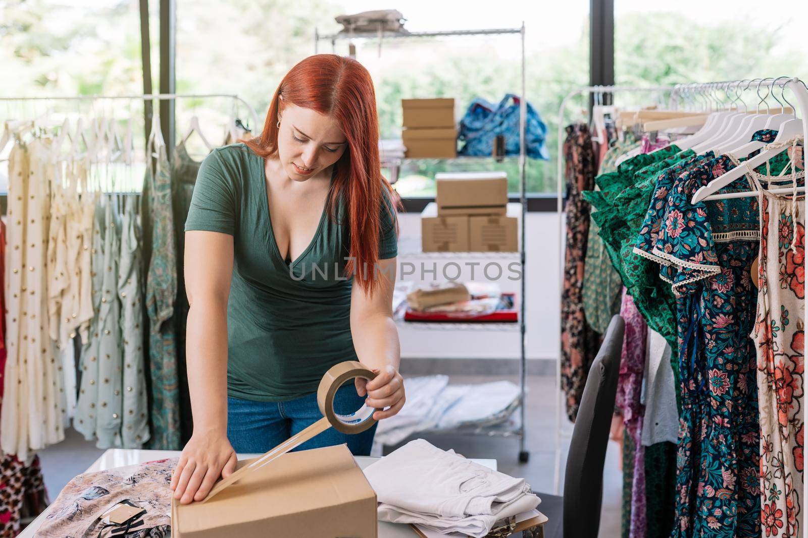 Young woman employee of an online clothing shop closes a cardboard box with adhesive tape, to send by courier to the buyer. Young woman preparing a package for a customer in the office. background with clothes racks and clothes.