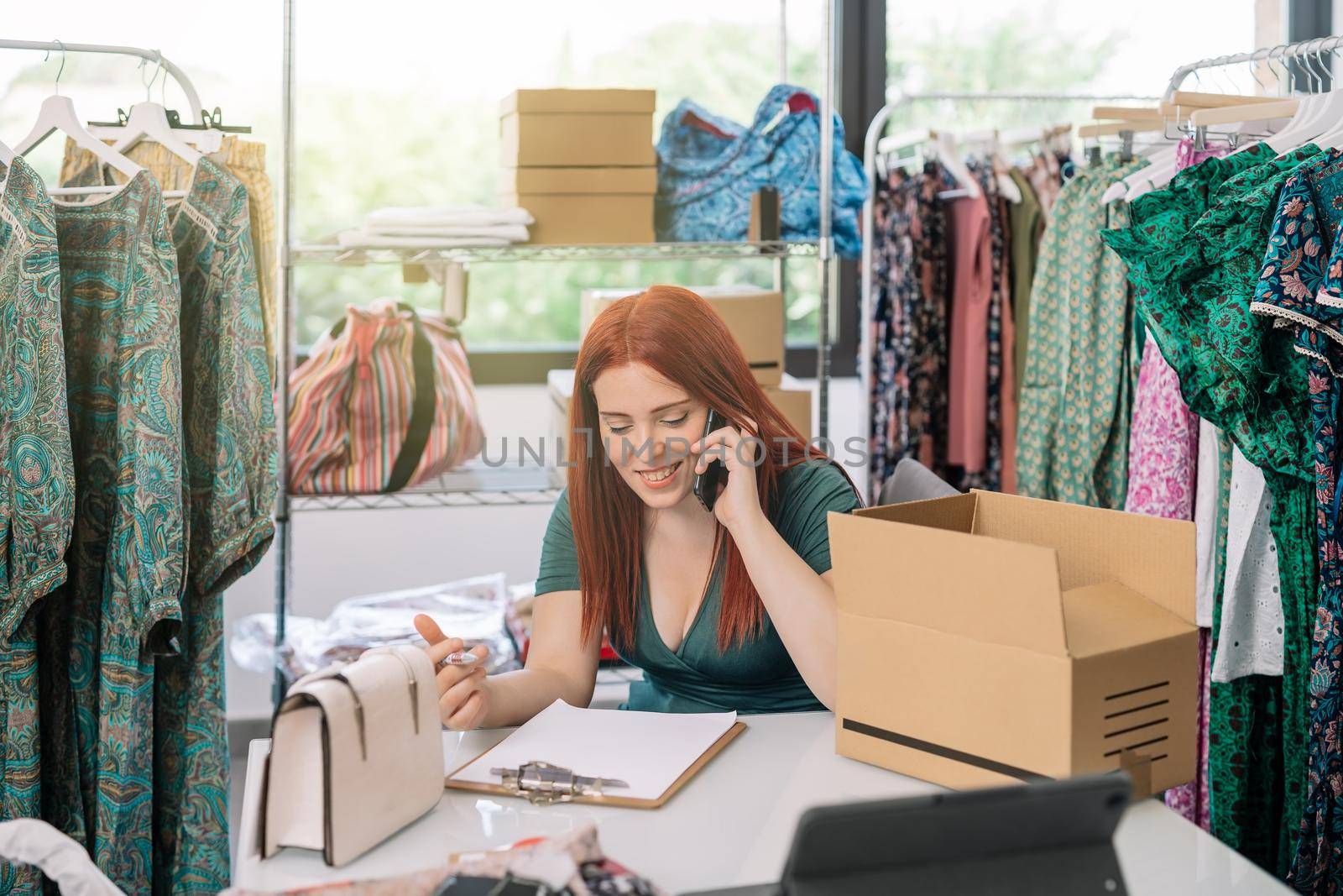 young woman businesswoman smiling, talking on her smartphone with her suppliers and customers in the warehouse of her clothing shop. work and business concept. by CatPhotography