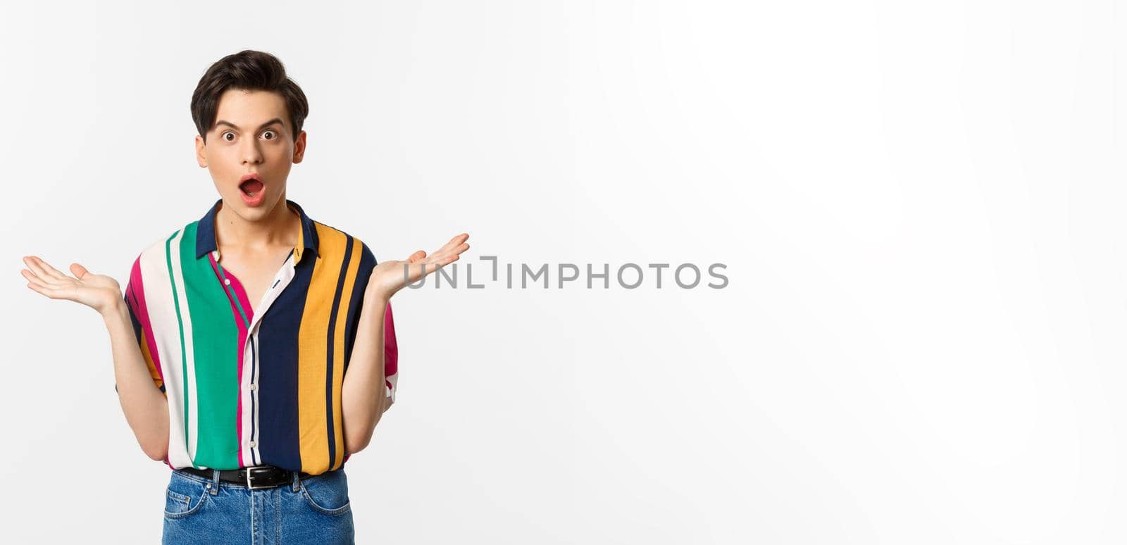 Image of surprised young male model staring at camera, spread hands sideways in complete disbelief, standing over white background.