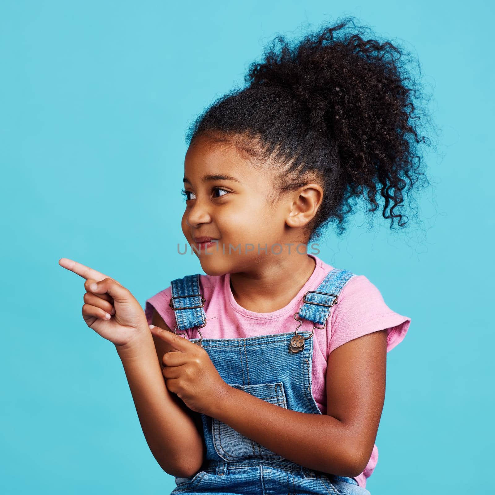 Im telling everyone. Shot of an adorable little girl pointing at something while standing against a blue background. by YuriArcurs