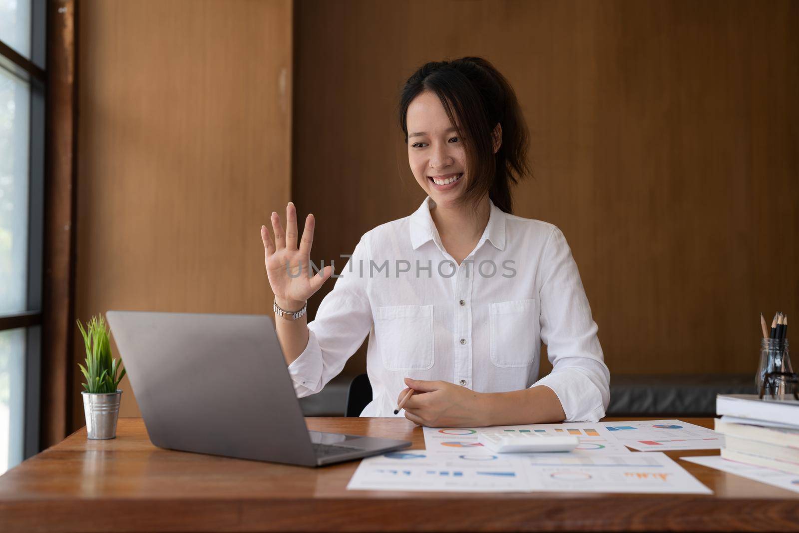 Young happy successful employee business asian woman waving hand sit work at workplace white desk talk by video call on laptop pc computer