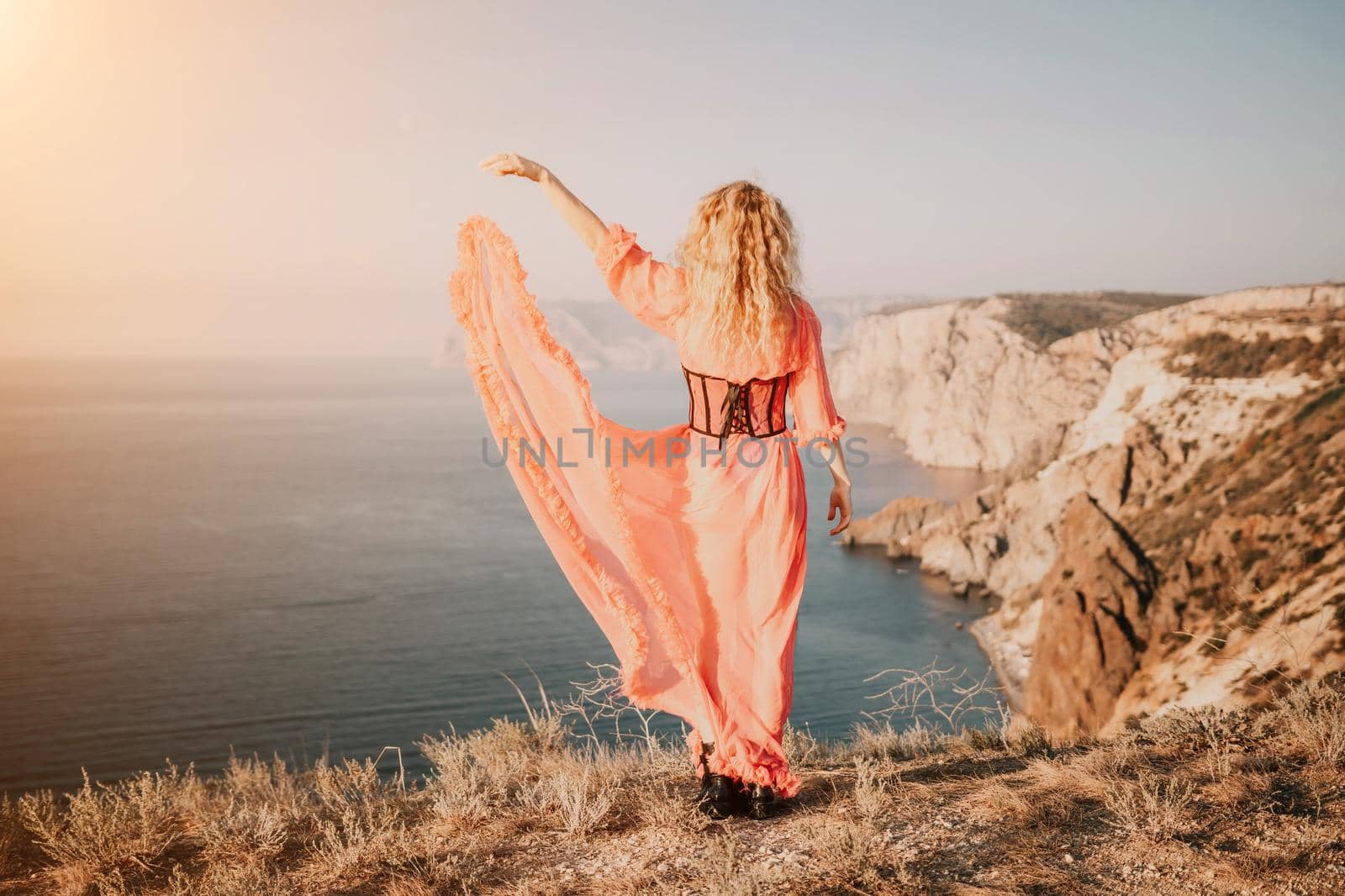 Beautiful young caucasian woman with curly blond hair and freckles. Cute redhead woman portrait in a pink long dress posing on a volcanic rock high above the sea during sunset. by panophotograph