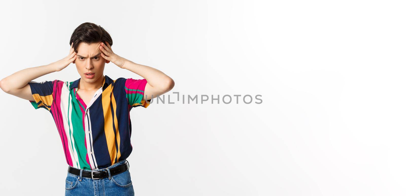 Anxious young gay man standing in panic, holding hands on head and staring down troubled, having a problem, standing over white background.