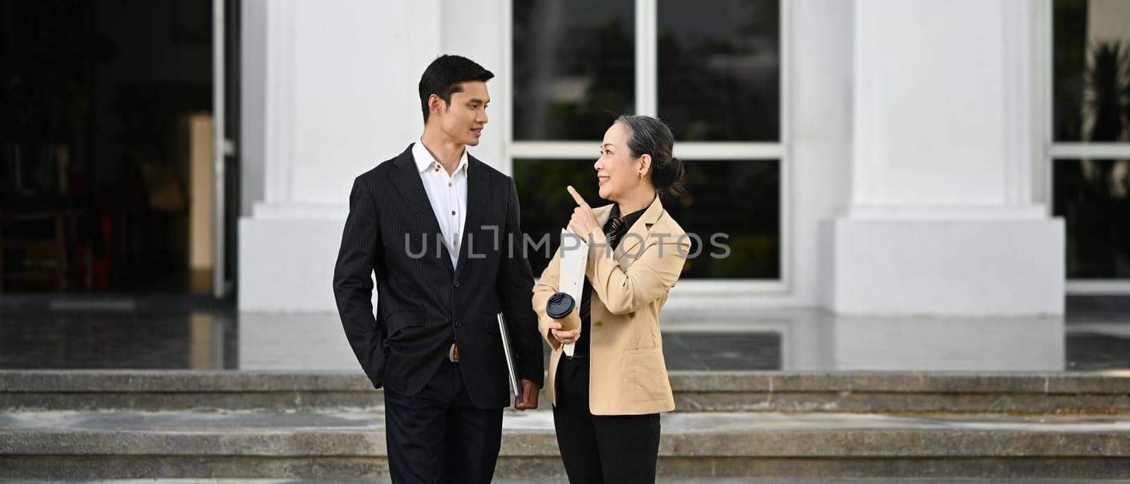 Two professional business people wearing formal suits talking and walking outside modern office building.