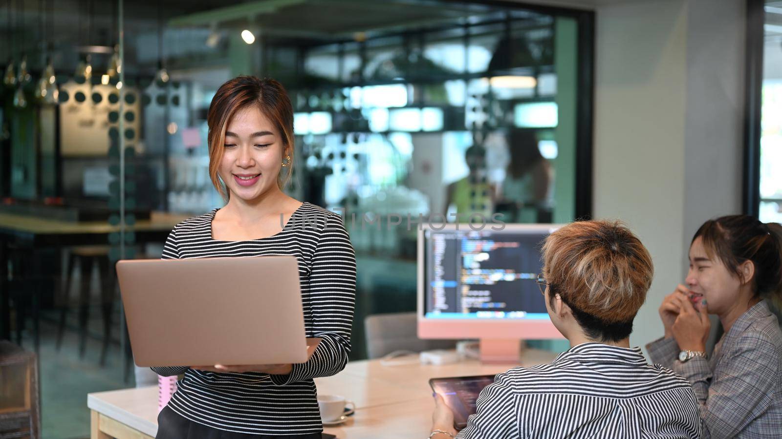 Asian female team leader standing in office and software developers team working in background.