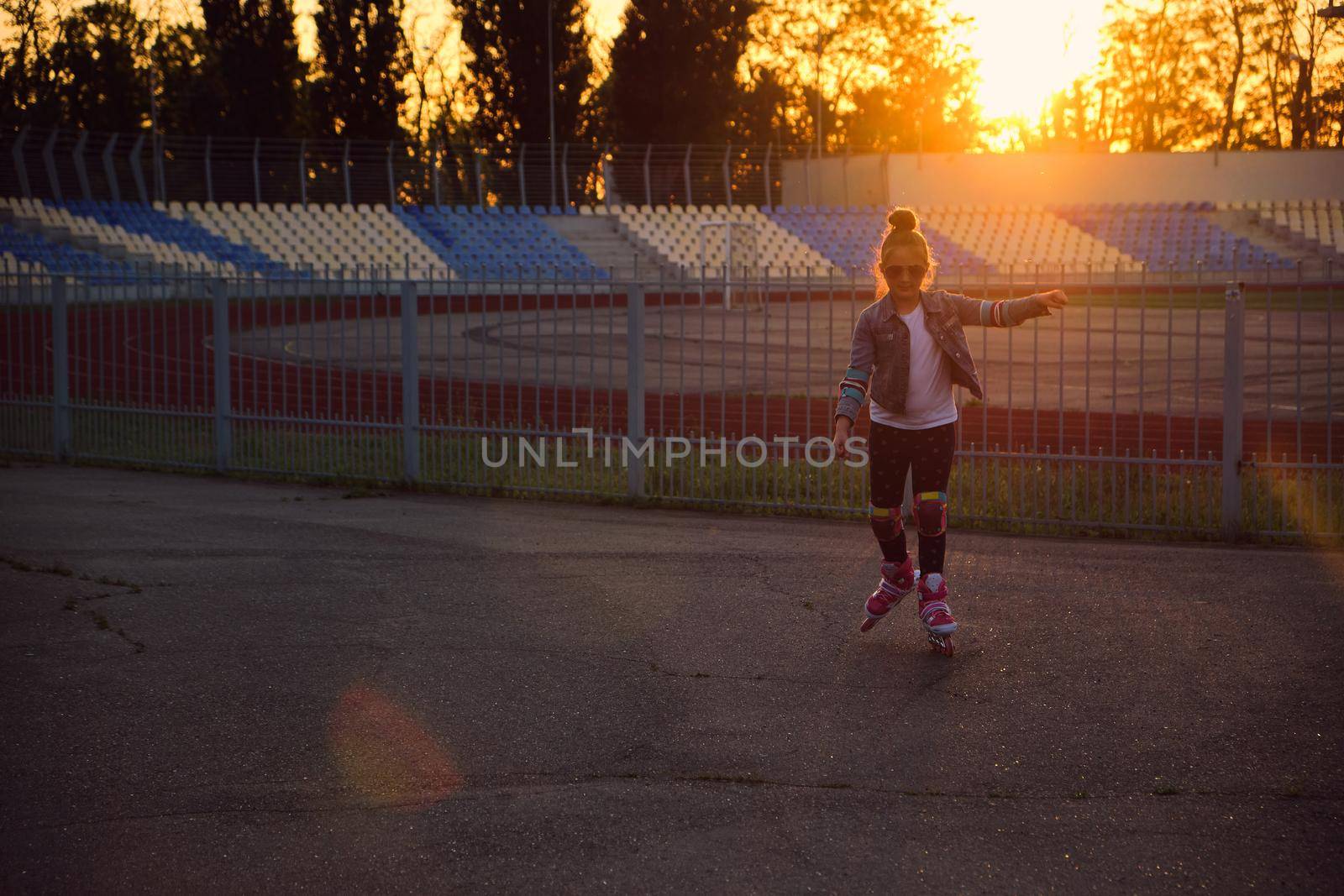 Little pretty happy funny girl on roller skates at stadium at sunset, learning to roller skate outdoors. Outdoor activity for children. Active sport for preschool kid. sun glare, selective focus