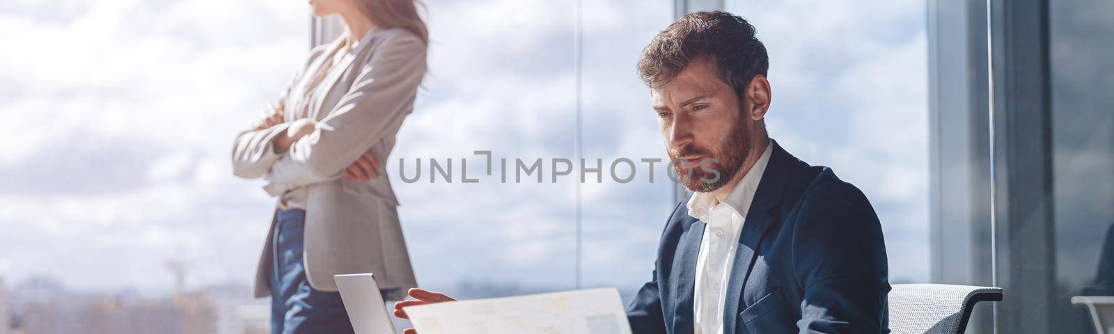 businessman analyzes work papers and charts while his colleague looks thoughtfully out the window by Yaroslav_astakhov