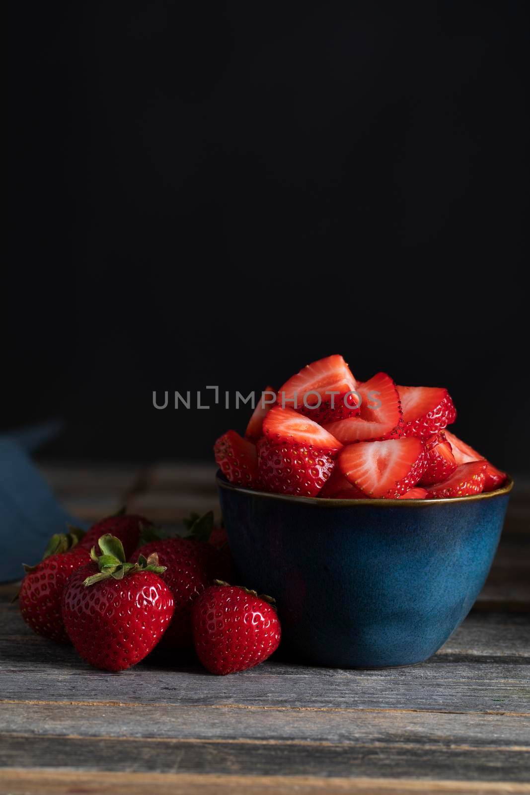 Fresh sliced strawberries in bowl with whole strawberries beside them, low key lighting and vertical orientation.