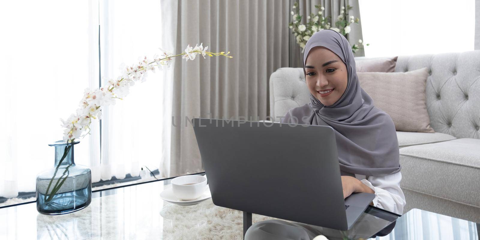 online education concept Smiling Arab woman wearing a headscarf on her laptop sits on the back of the sofa at home. enjoy distance learning copy space.