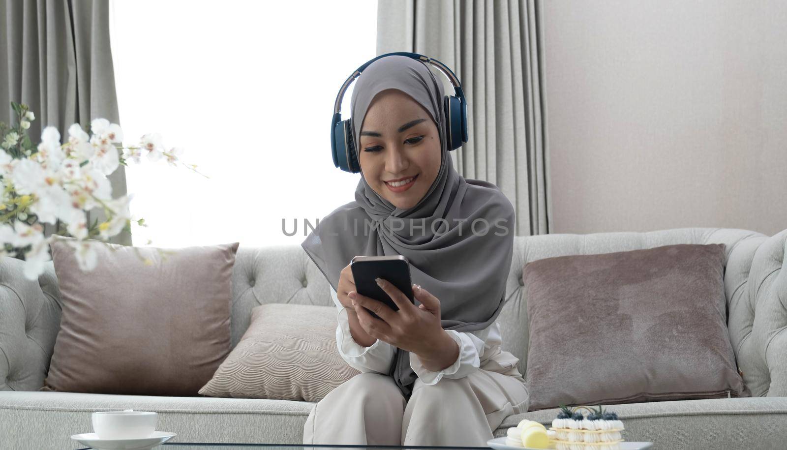 Modern leisure. Muslim girl wearing hijab and using smartphone for listening music in wireless headphones at home, empty space by wichayada