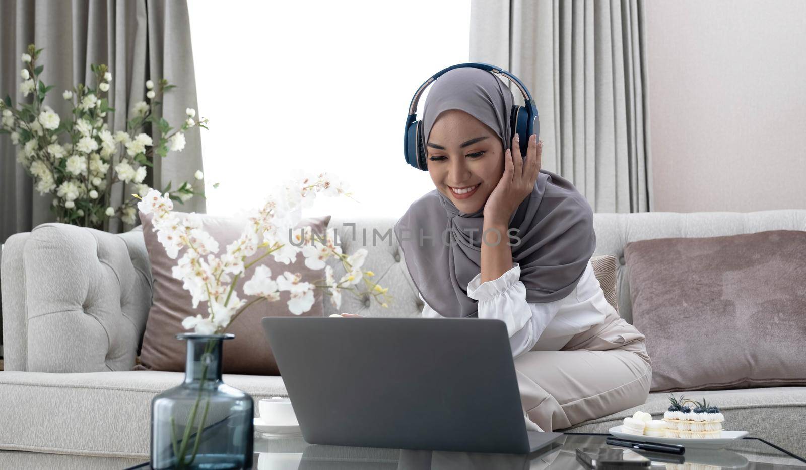 Playlist for relaxation Portrait of a calm Muslim woman in hijab listening to music with her laptop and wireless headphones at home. A smiling Islamic woman sits with her eyes closed in a comfortable chair..