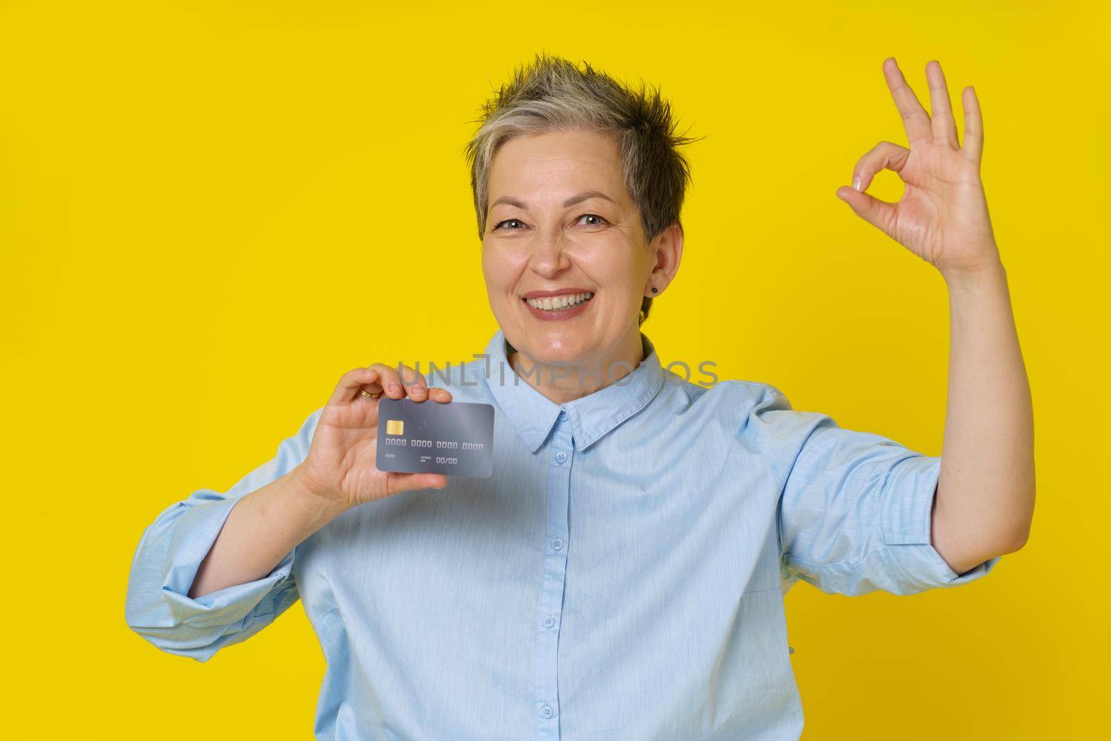 Holding credit, credit card grey haired woman gesturing OK with other hand making online payment or shopping. Pretty mature woman in blue shirt isolated on yellow background. Banking concept by LipikStockMedia