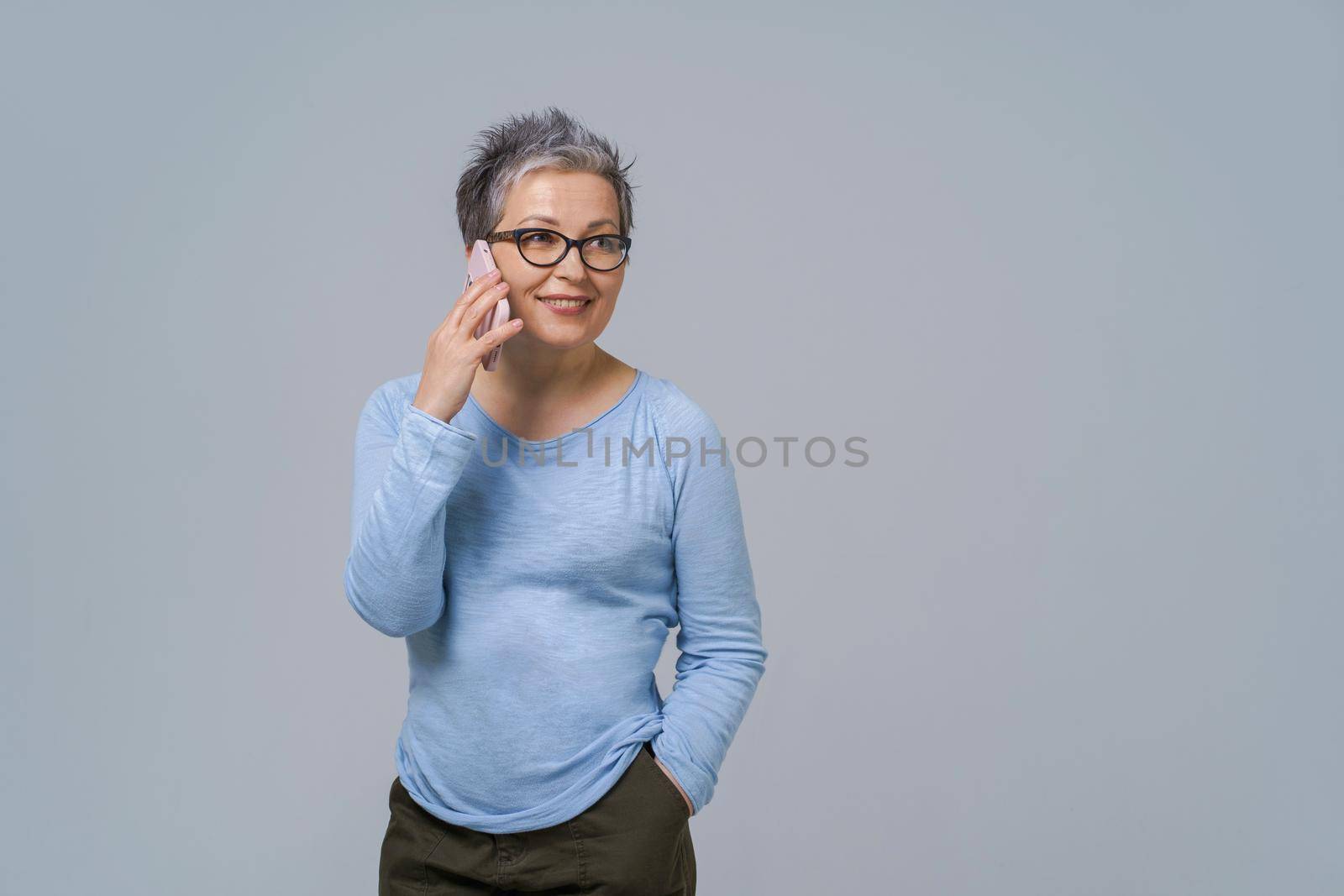 Talking on the phone mature grey hair woman in glasses turn sideways holding smartphone in her hand wearing blue blouse and black skirt. Pretty woman in blue shirt isolated on white background by LipikStockMedia