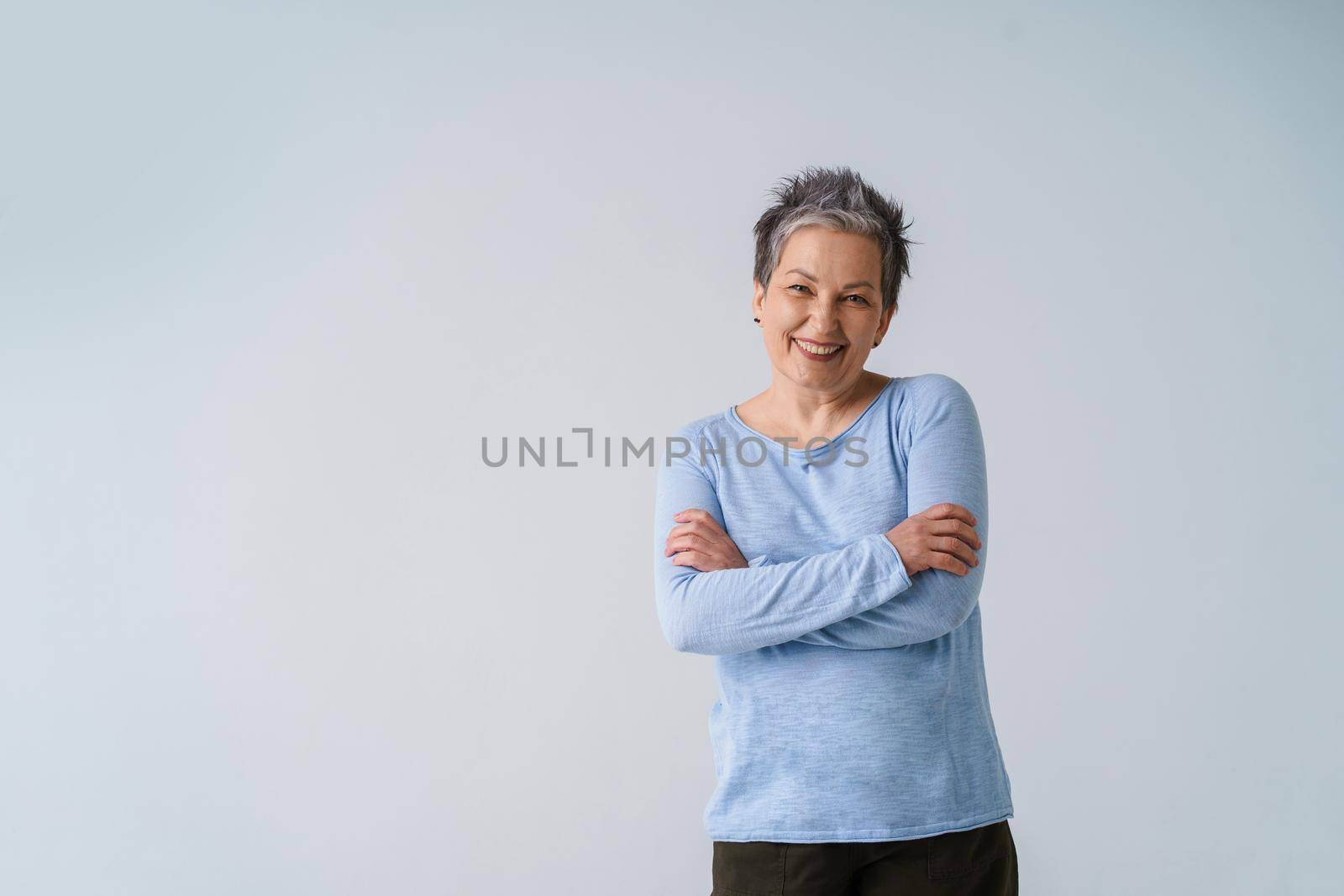 Charming positive emotions mature grey hair woman posing with hands folded looking at camera wearing blue blouse, copy space isolated on white background. Healthcare, aged beauty concept.