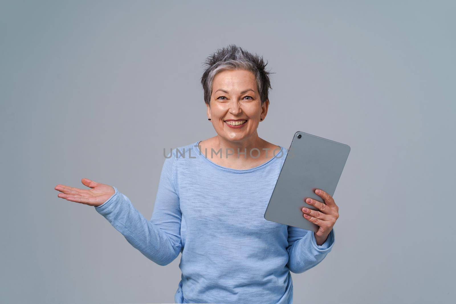 Happy middle age woman with grey hair and tablet pc in hands browsing, working, shopping online. Pretty woman in 50s wearing blue blouse isolated on grey. Mature people and technologies.