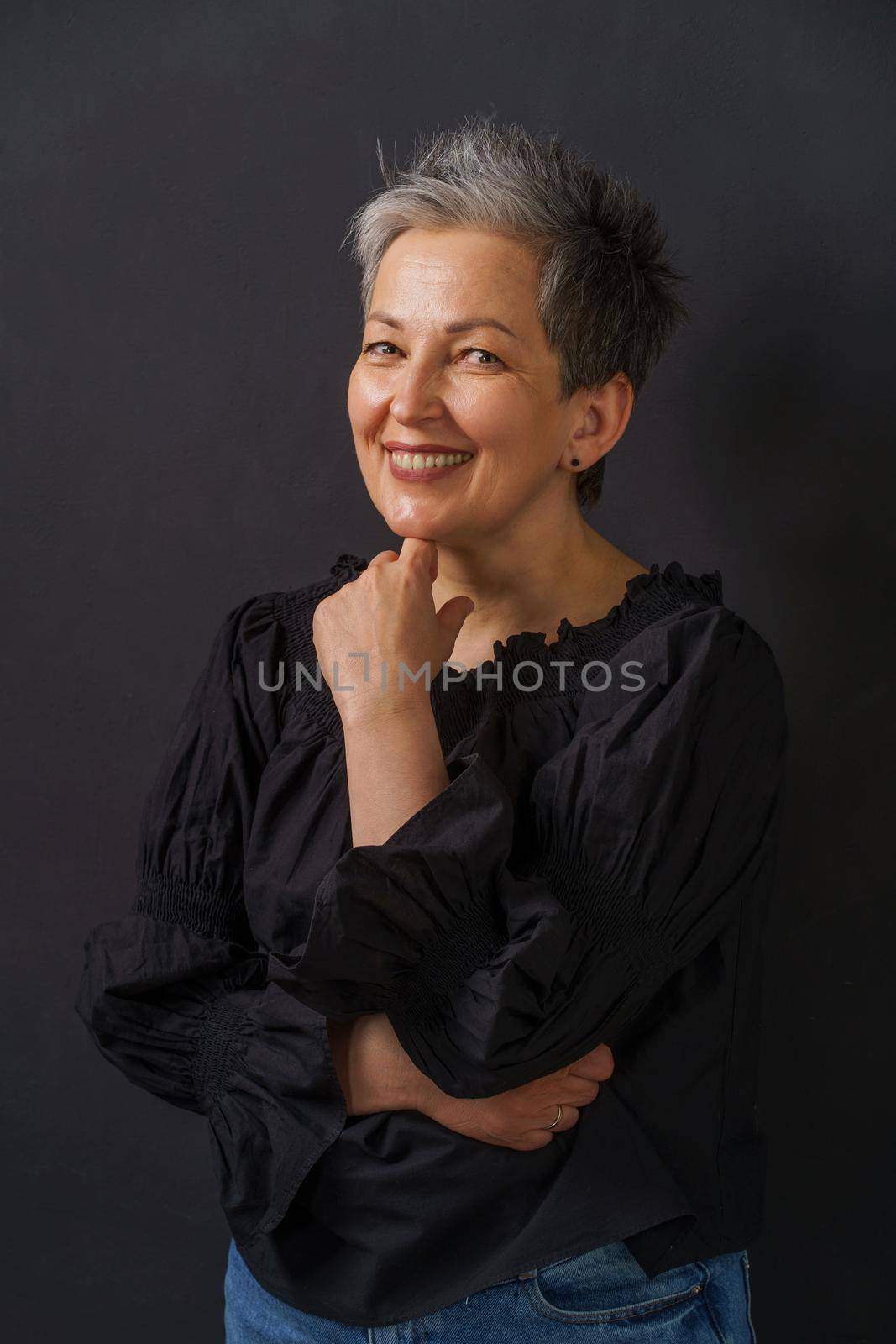 Mature businesswoman with grey hair smiling posing leaned chin on folded hands isolated on black background. Graying elegant woman with short hair. Image of successful mature woman. Aged beauty.