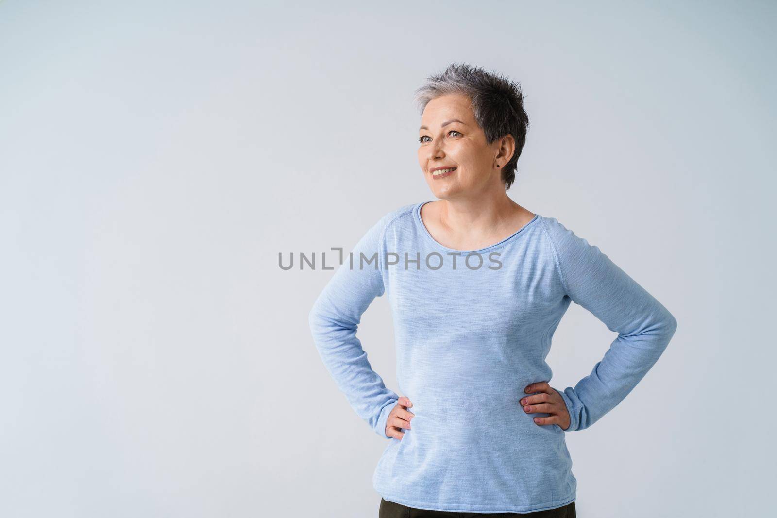 Charming mature silver, grey hair woman posing with hands on her hips looking sideways away wearing blue blouse, isolated on white background. Healthcare, aged beauty concept. Copy space.