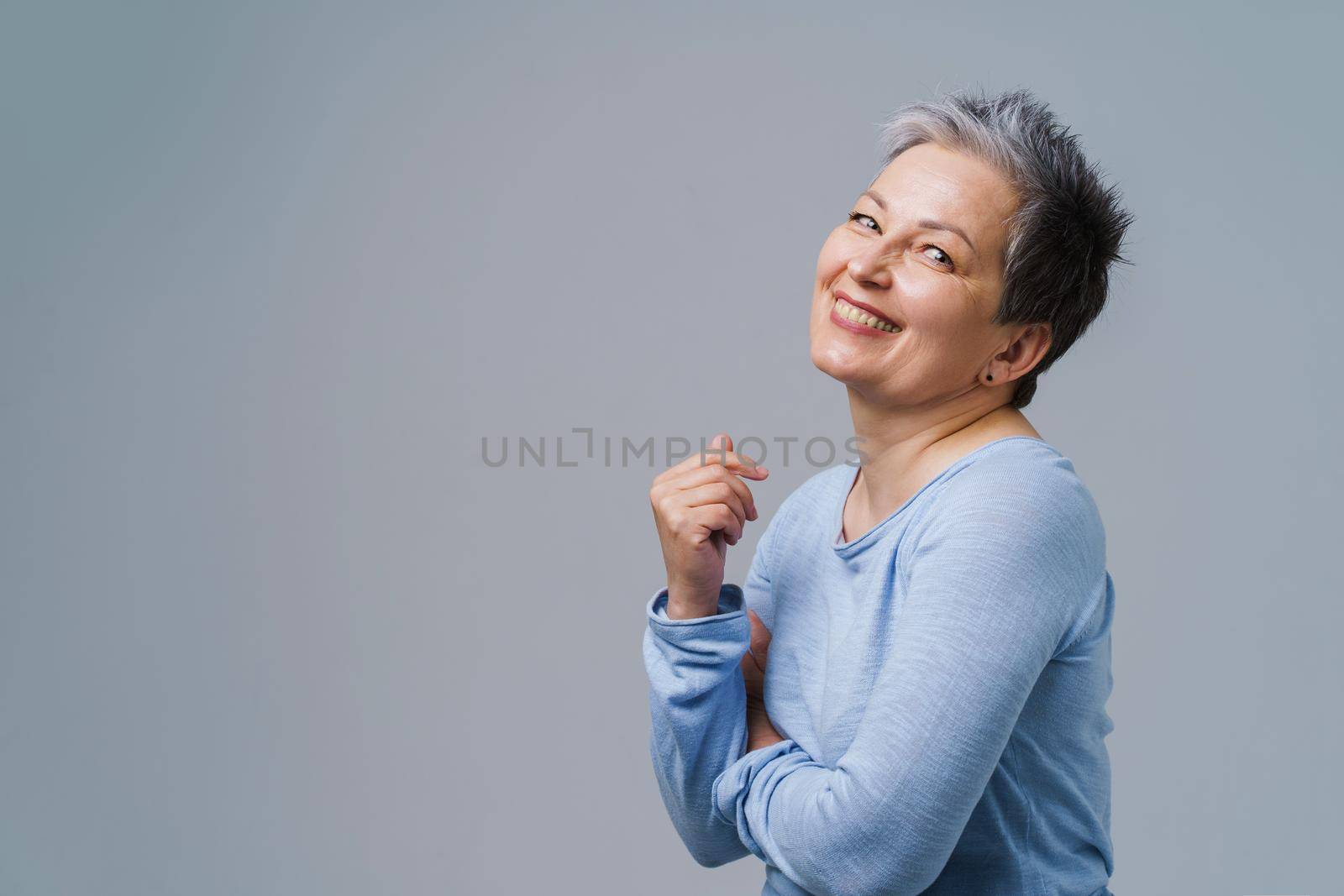 Positive emotions mature grey hair woman posing with hands folded looking at camera wearing blue blouse, copy space on left isolated on white background. Healthcare, aged beauty concept.