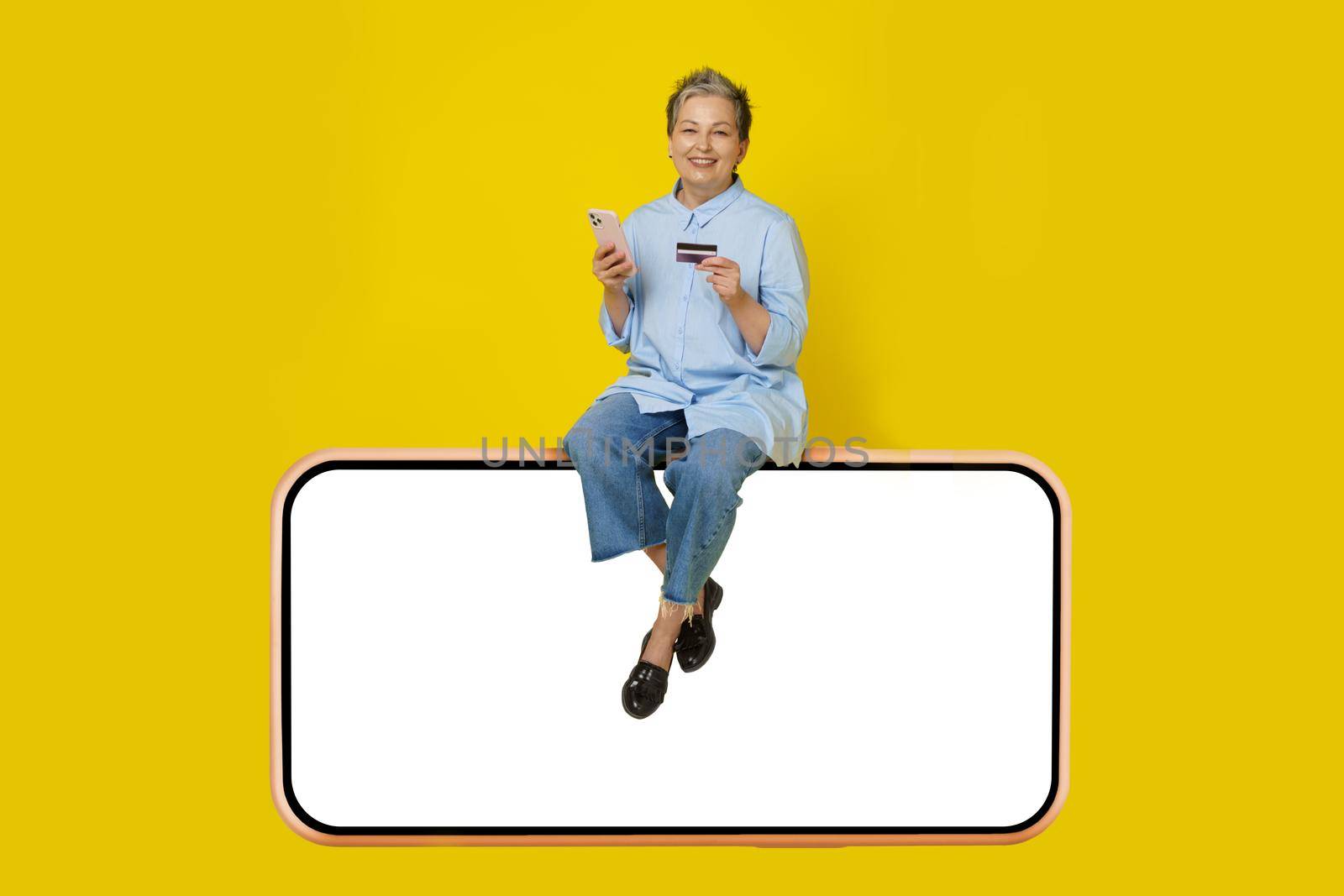 Silver, grey haired woman with phone and debit, credit card in hand making online payment or shopping sitting on a huge, giant smartphone, wearing blue shirt and jeans isolated on yellow background by LipikStockMedia