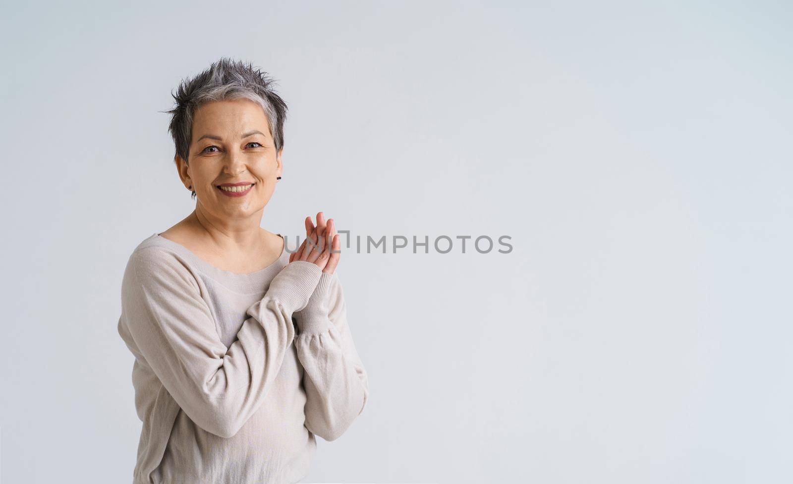 Mature grey hair woman posing with hands folded smiling looking at camera wearing white blouse, copy space on right isolated on white background. Healthcare concept. Aged beauty concept.