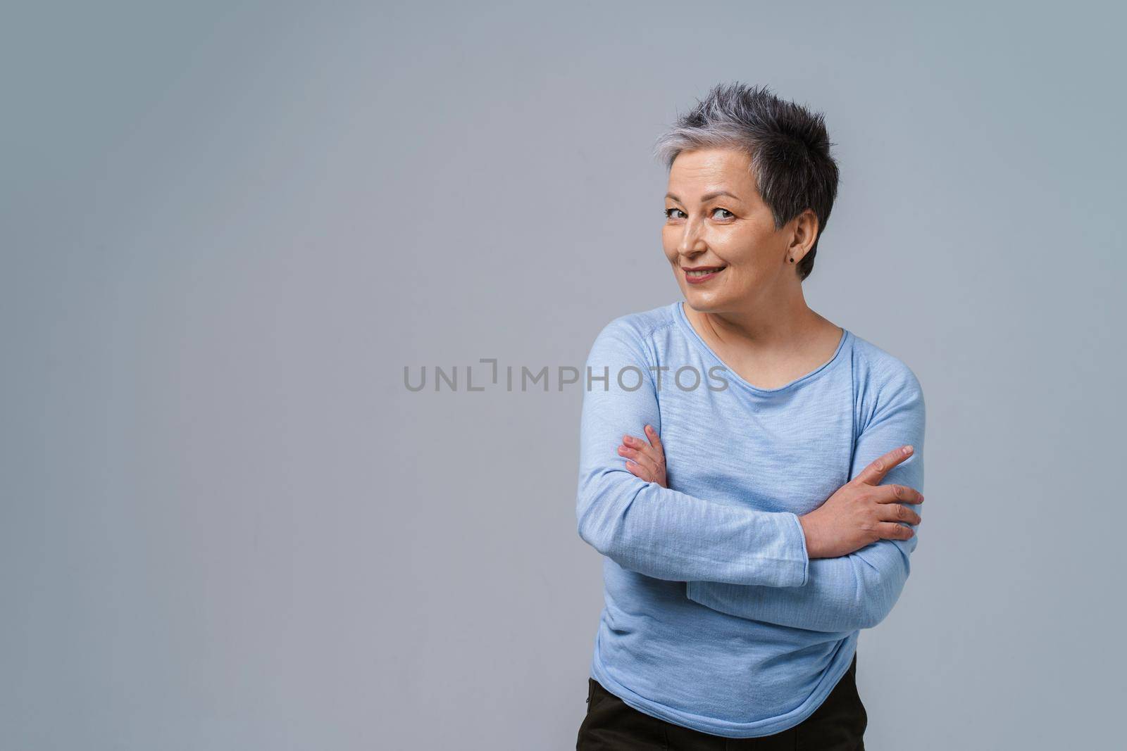 Challenging look positive emotions mature grey hair woman posing with hands folded looking at camera wearing blue blouse, isolated on white background. Healthcare, aged beauty concept. Copy space by LipikStockMedia