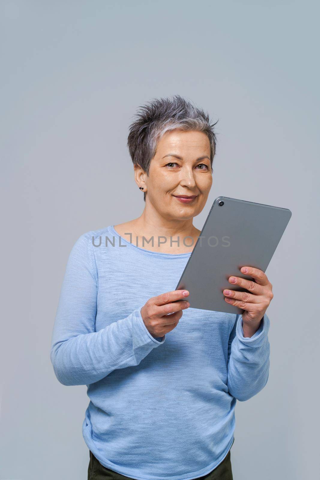 Grey haired woman with tablet pc in hands working online. Pretty woman in 50s wearing blue blouse isolated on grey. Mature people and technologies.