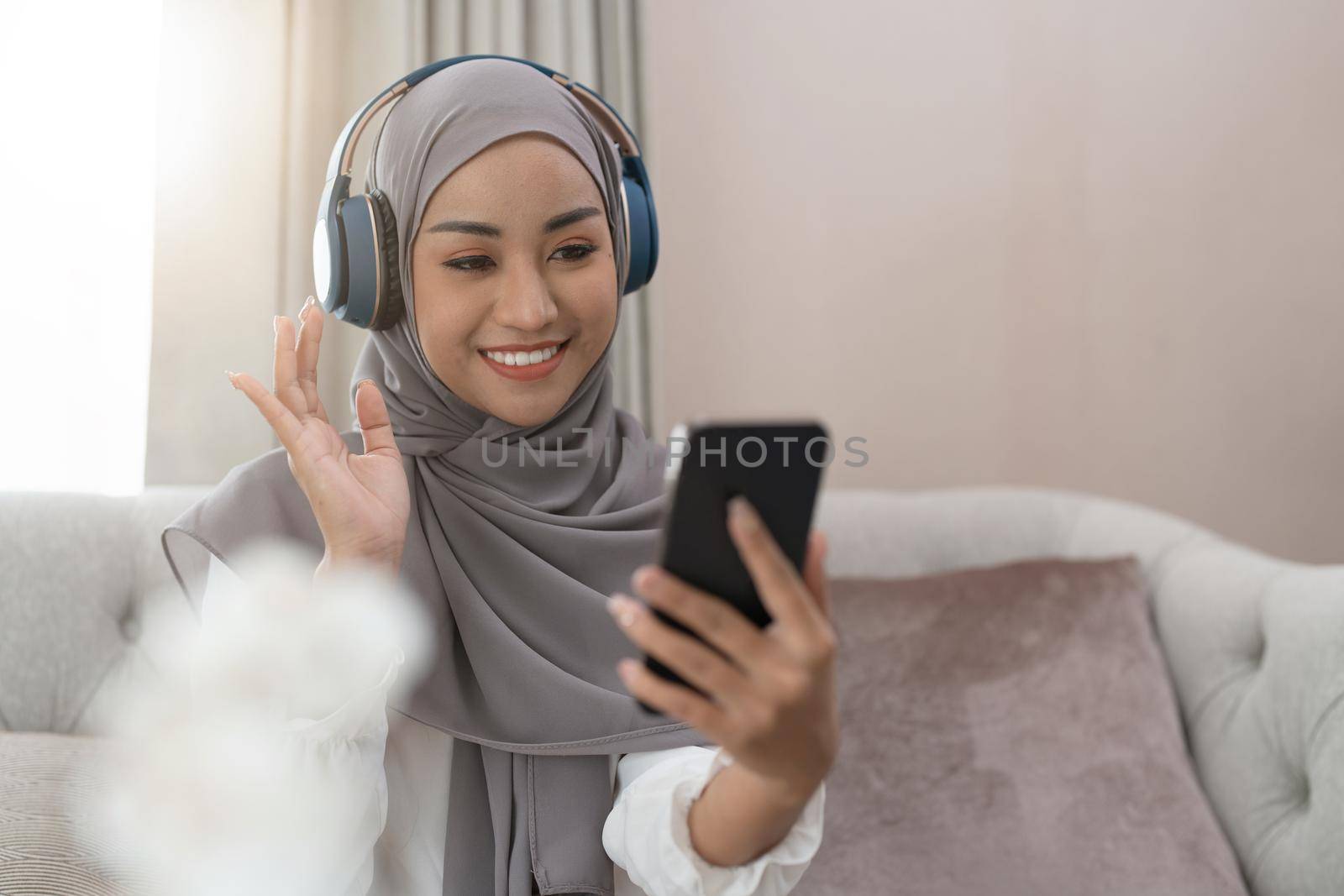 Muslim woman sitting on sofa and video conference calling on mobile phone. Muslim asian woman video call. by nateemee