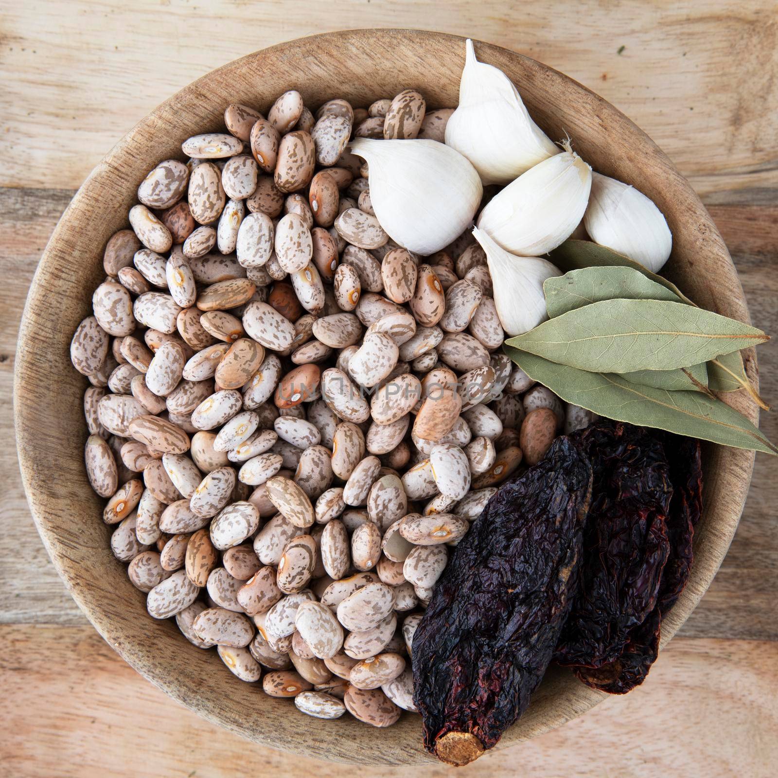 Flat lay of wooden bowl full of pinto beans, garlic, bay leaves and dried poblano peppers.