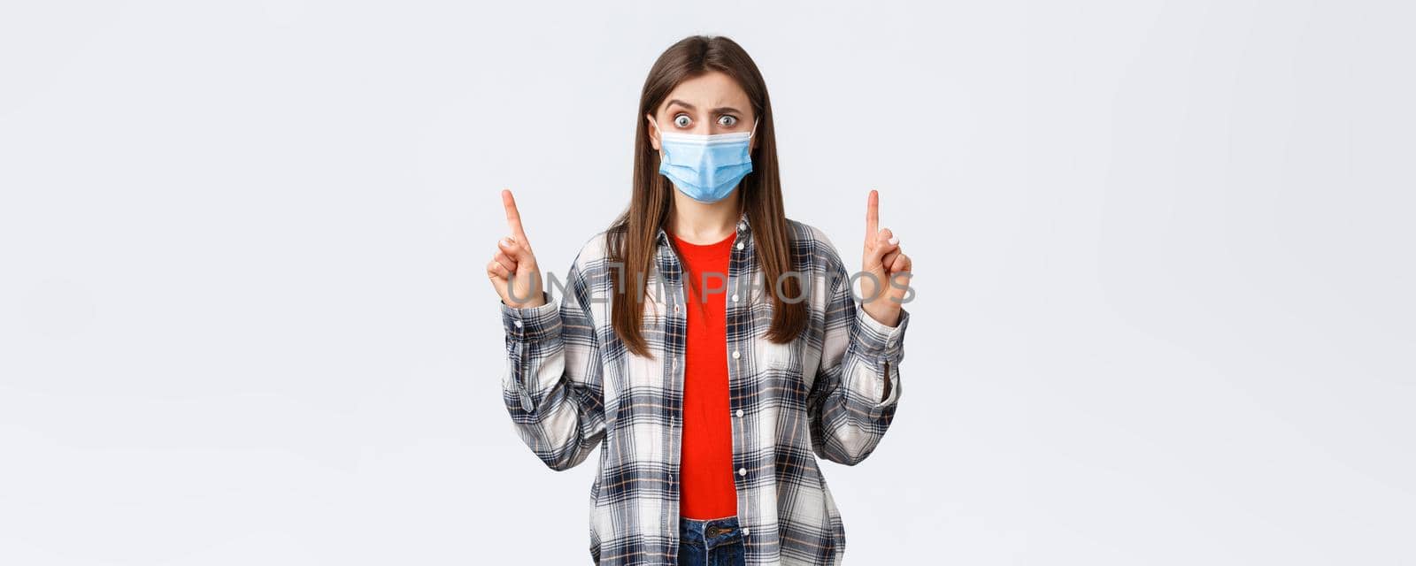 Coronavirus outbreak, leisure on quarantine, social distancing and emotions concept. Concerned and puzzled young woman in medical mask pointing fingers up at strange, confusing promo by Benzoix