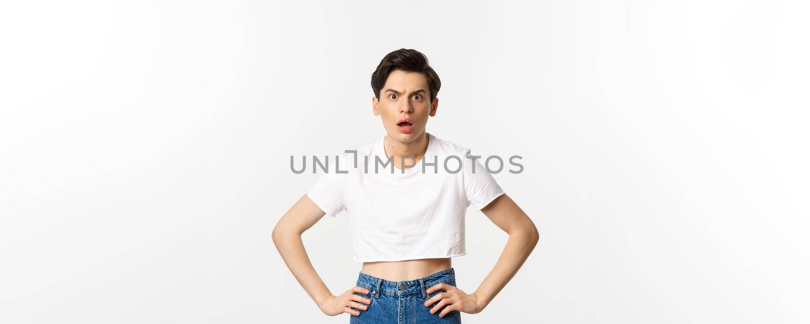 Shocked and confused gay man staring at camera startled, wearing crop top, holding hands on waist, standing over white background.