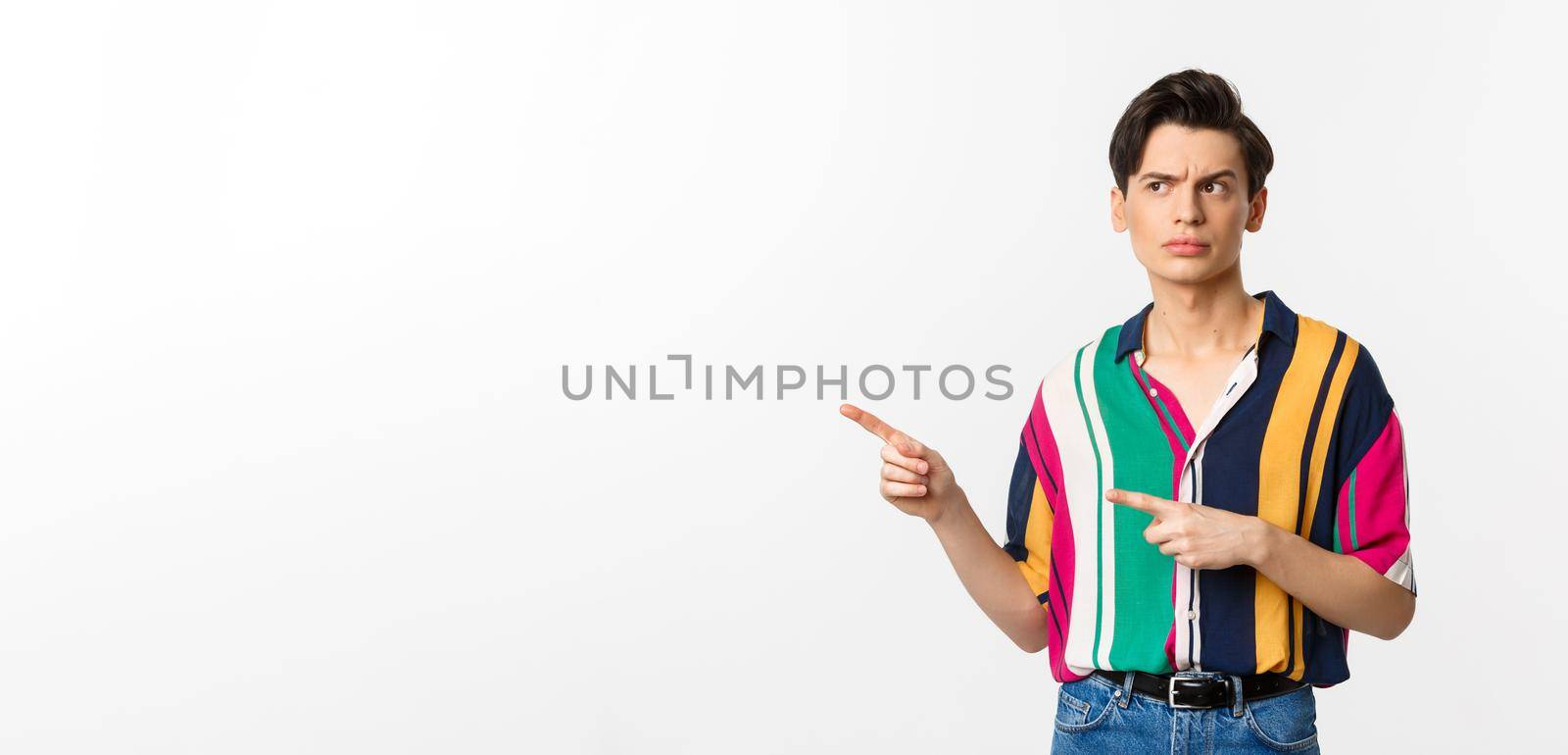 Suspicious young man looking and pointing fingers left with doubtful face, standing unsure in summer shirt, white background by Benzoix