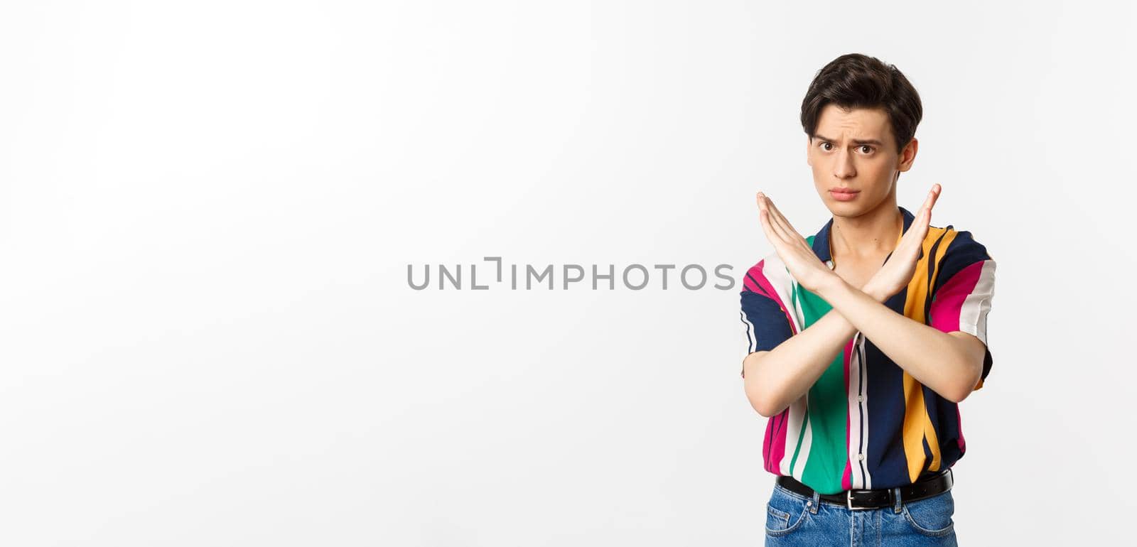 Disappointed young queer man telling no, showing stop gesture and forbid something bad, standing over white background.